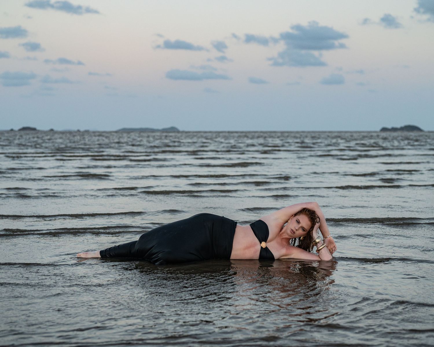 Woman in a black top and skirt lies on her side in shallow ocean water at dusk, with distant islands on the horizon.