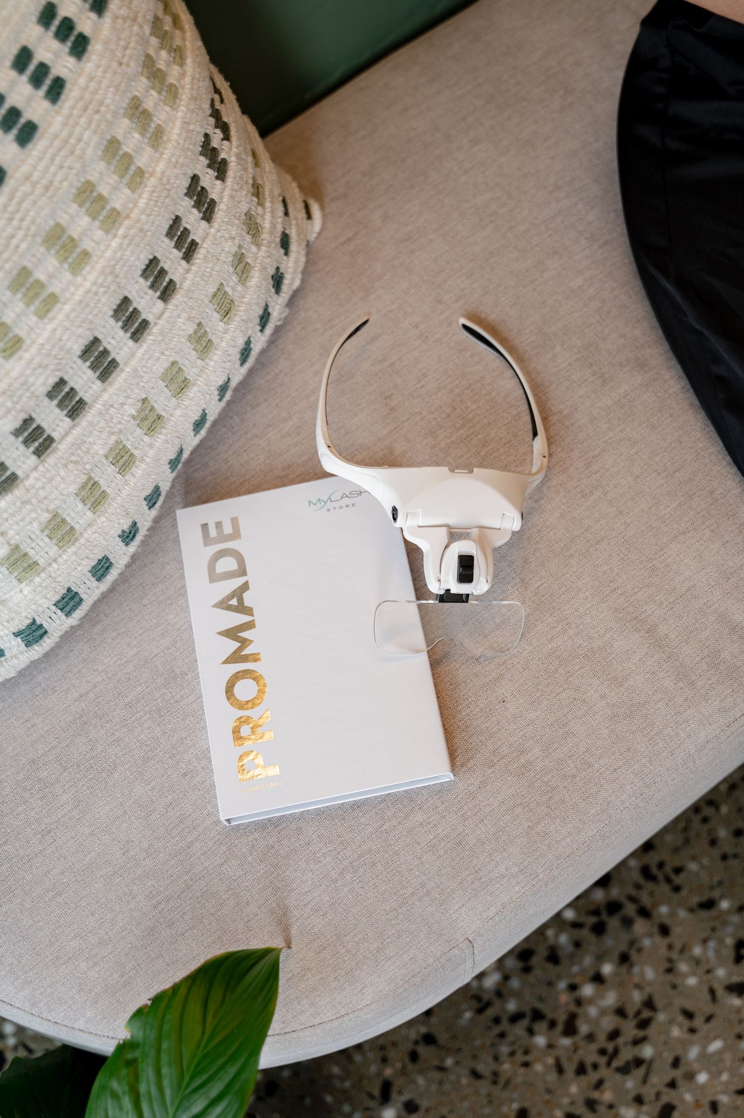 White magnifying headband glasses rest on a Promade booklet on a gray cushioned seat beside a patterned pillow and green plant leaf.