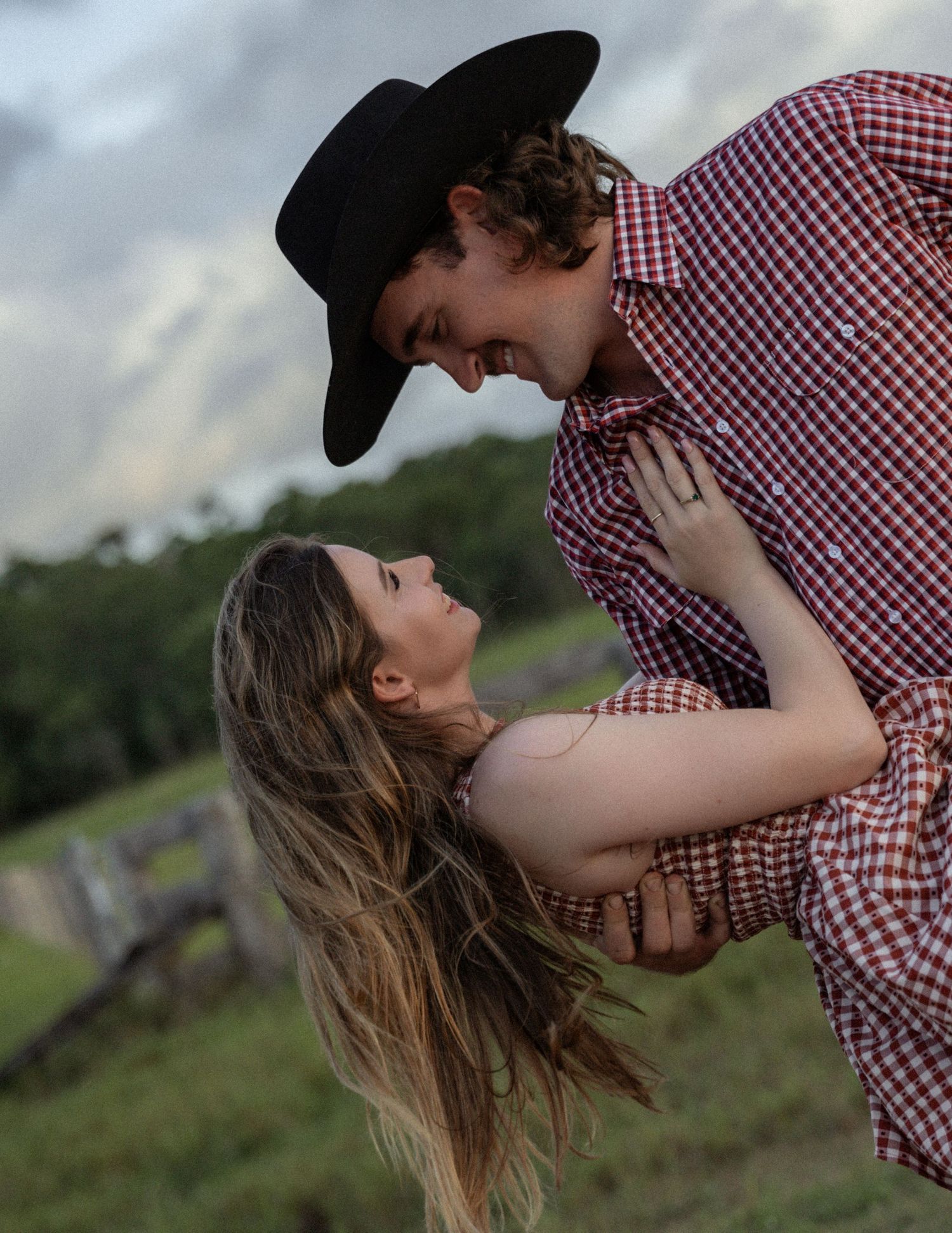 Smiling cowboy leans in close to a woman with long hair held in a romantic dip beneath a cloudy sky on a country farm.