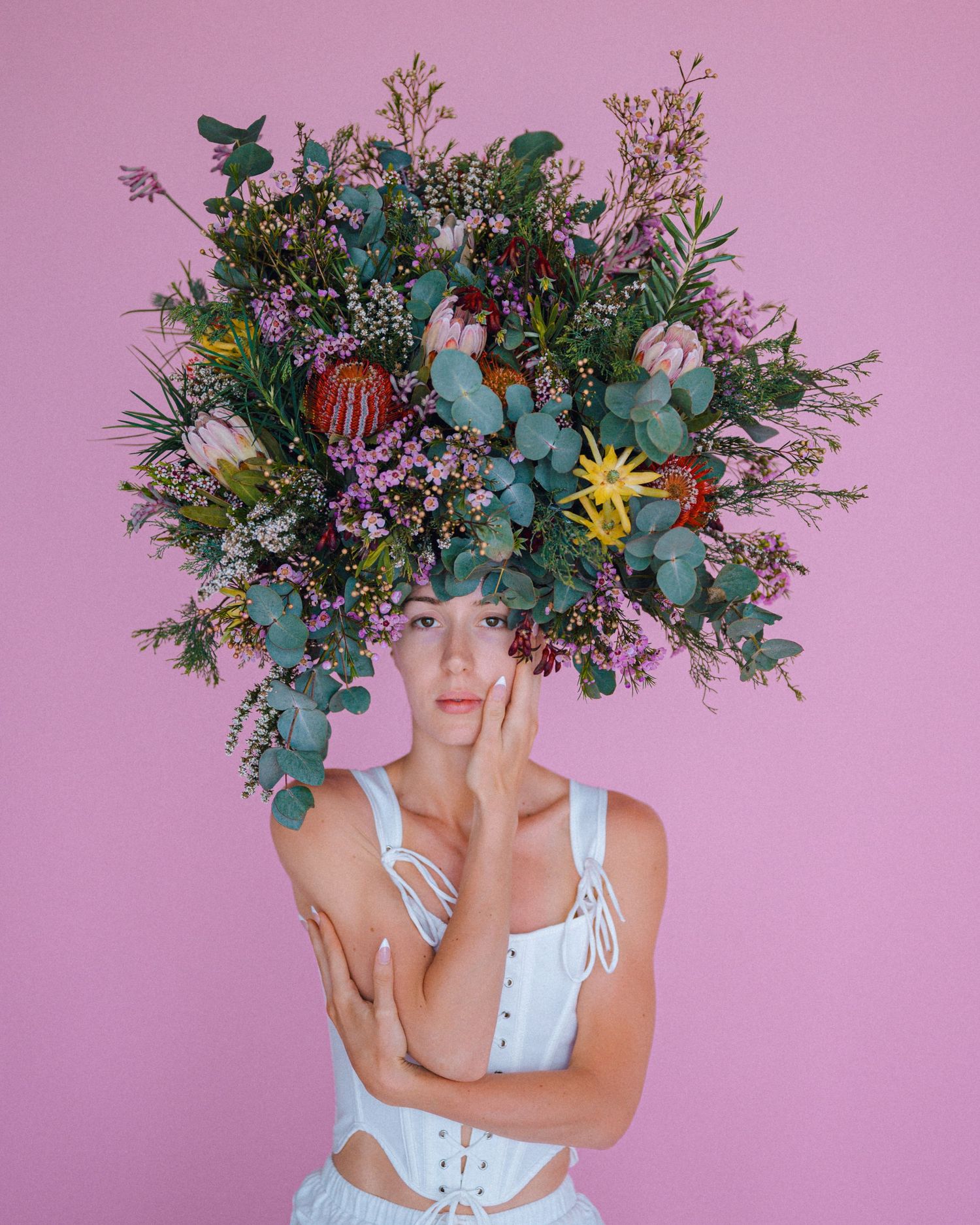 Model wearing an oversized floral headpiece of eucalyptus, proteas and wildflowers with a white corset top against a pink background.