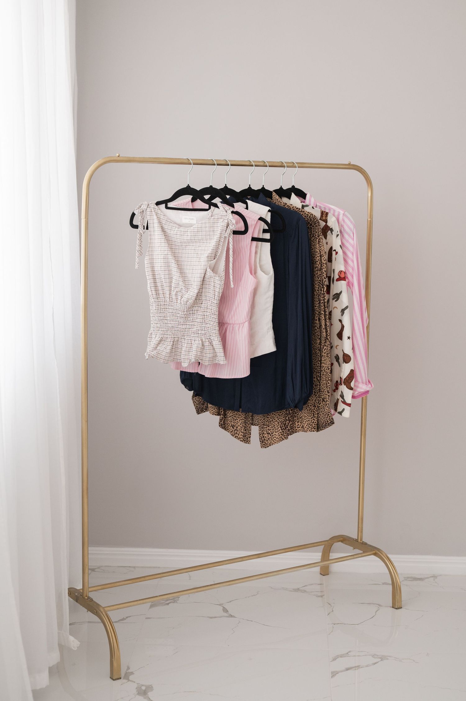 Gold clothing rack holding blouses, skirts and dresses in pink, navy, and floral prints beside sheer white curtains.