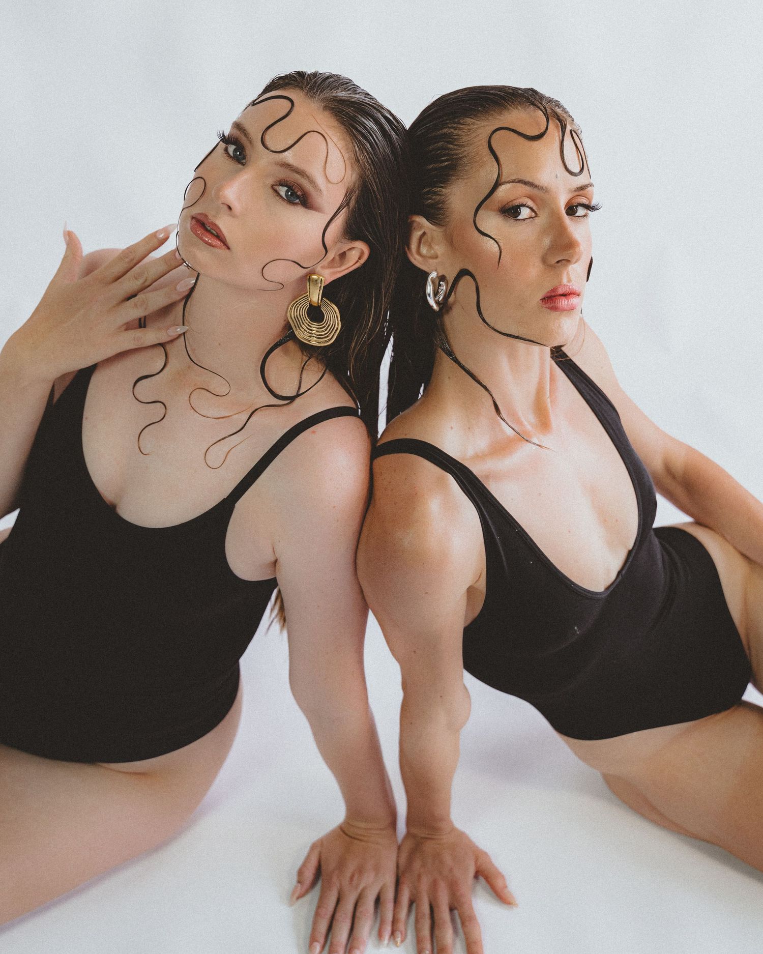 Two women in black bodysuits pose together against a white background, wearing gold earrings and swirling face line art.
