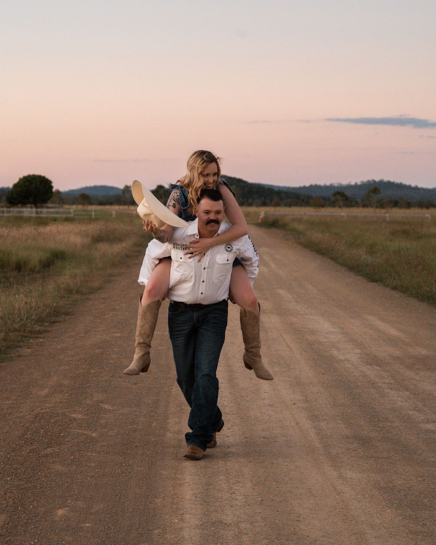 Man in a white shirt gives a woman a piggyback ride along a dirt road in the countryside at pink sunset.