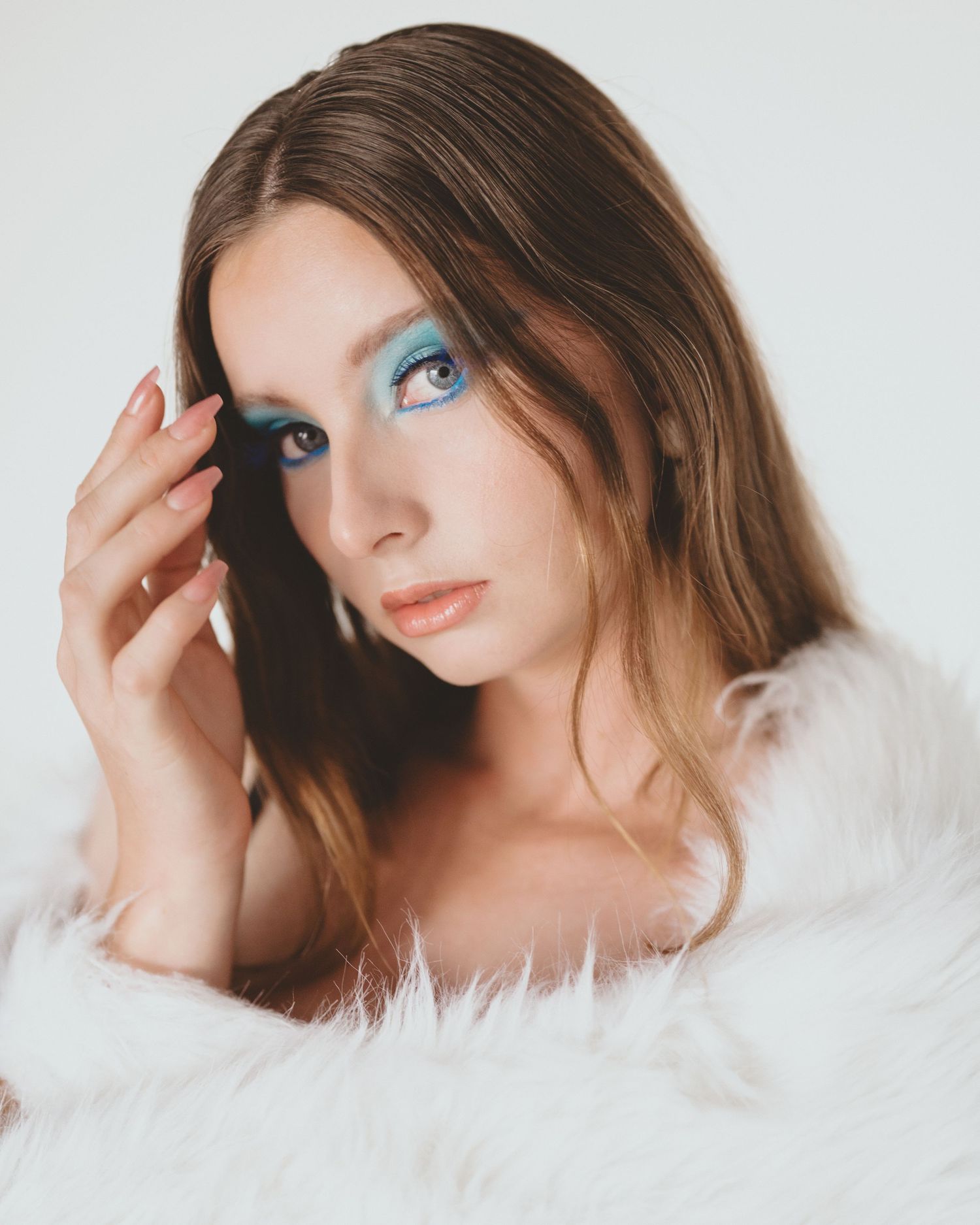 Beauty portrait of a woman with slicked-back hair, turquoise eye makeup and a white fur wrap, one hand raised near her face.