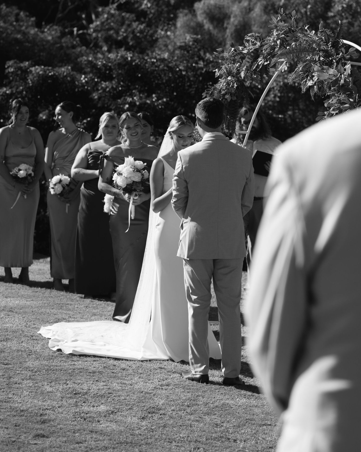 Black and white photo of a bride and groom exchanging vows outdoors while bridesmaids with bouquets look on.