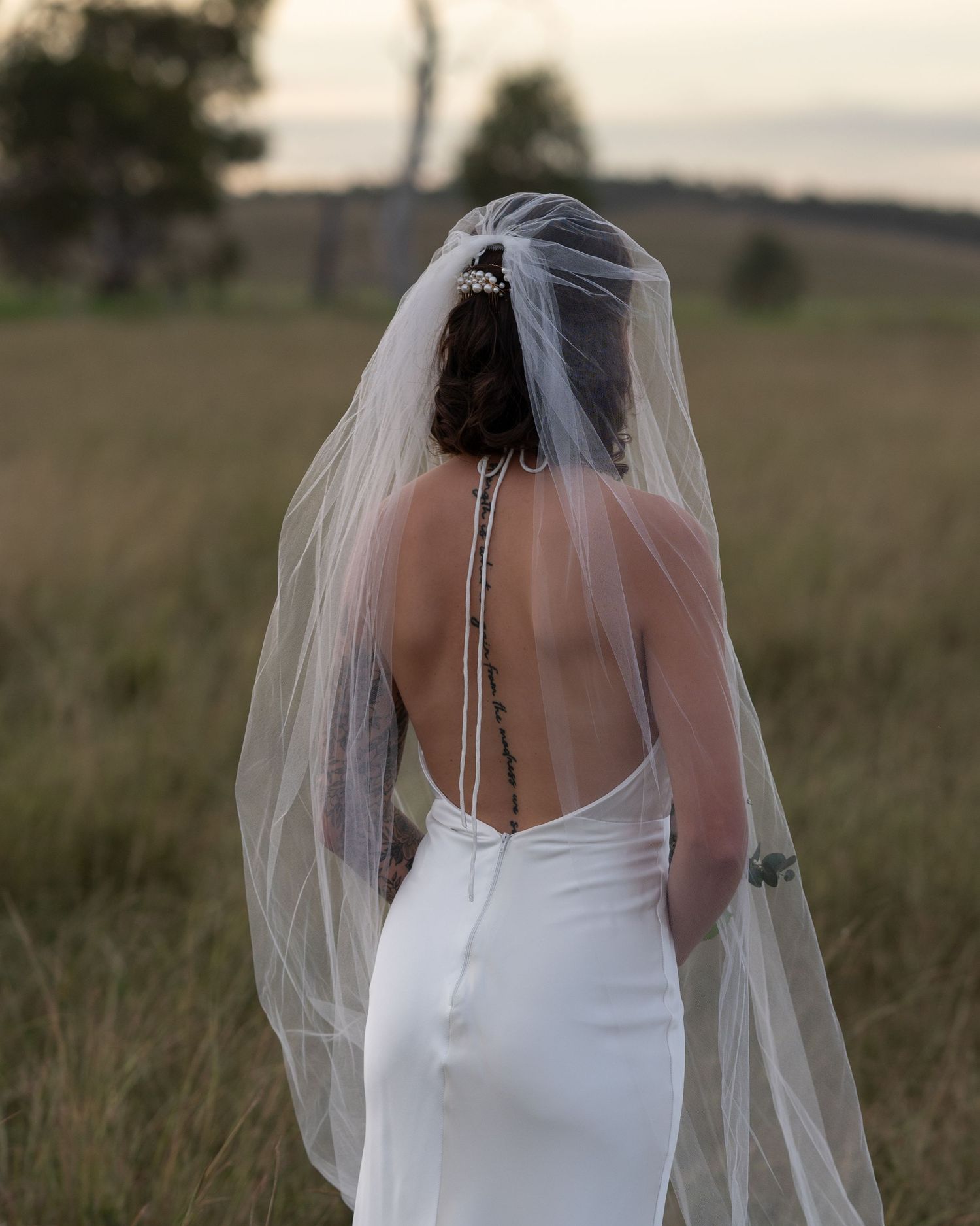 Bride seen from behind in a low-back white wedding dress and sheer veil, gazing across a grassy field.