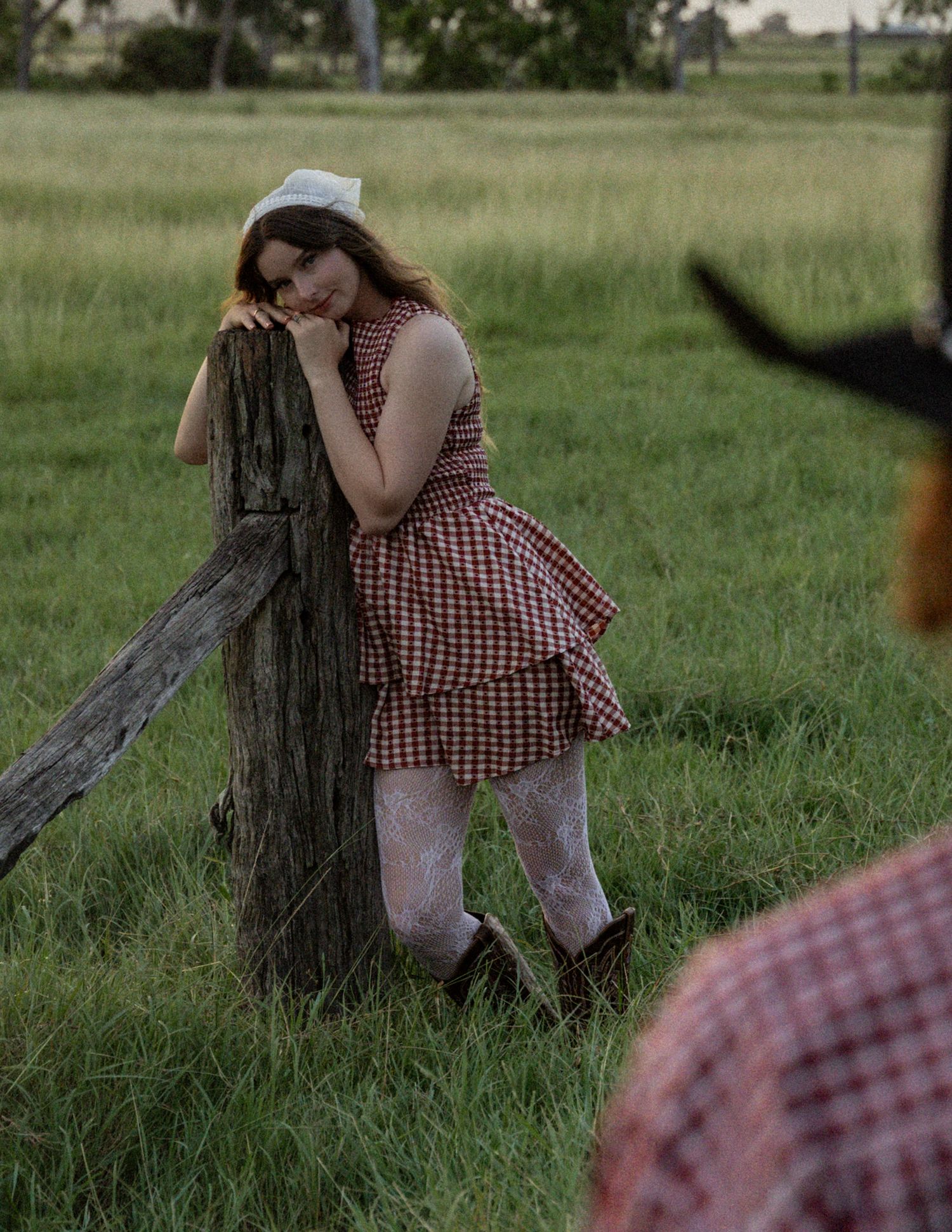 Woman in a red gingham dress, lace tights and cowboy boots leans dreamily on a rustic wooden fence post in a green pasture.