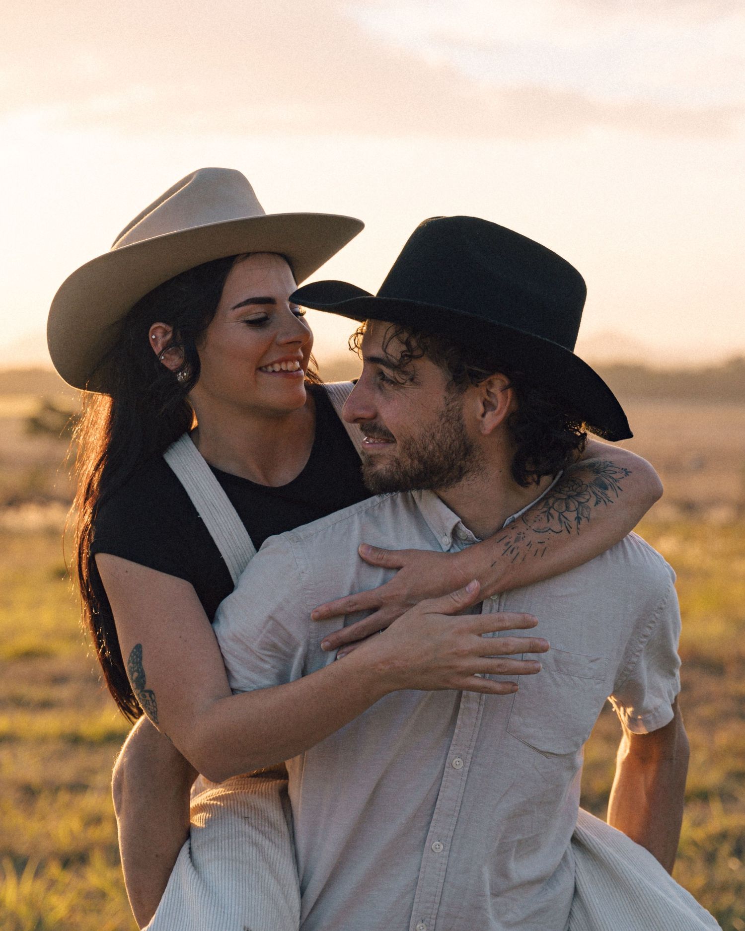 Smiling woman in a tan hat hugs a bearded man in a black hat from behind in a golden grassy field at sunset.