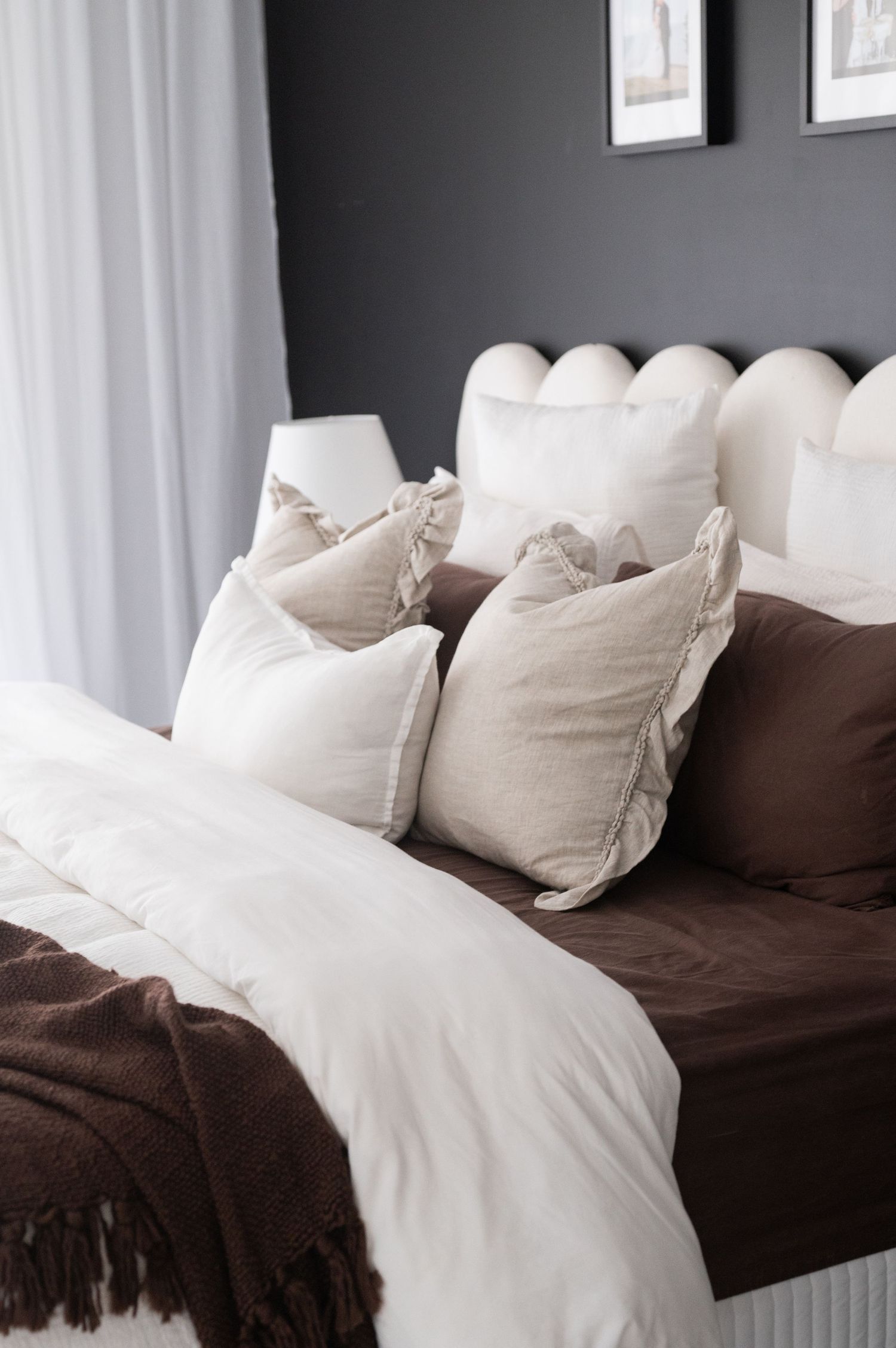 Neatly made bed with cream and chocolate brown linen, ruffled cushions, a knit throw and a scalloped headboard.