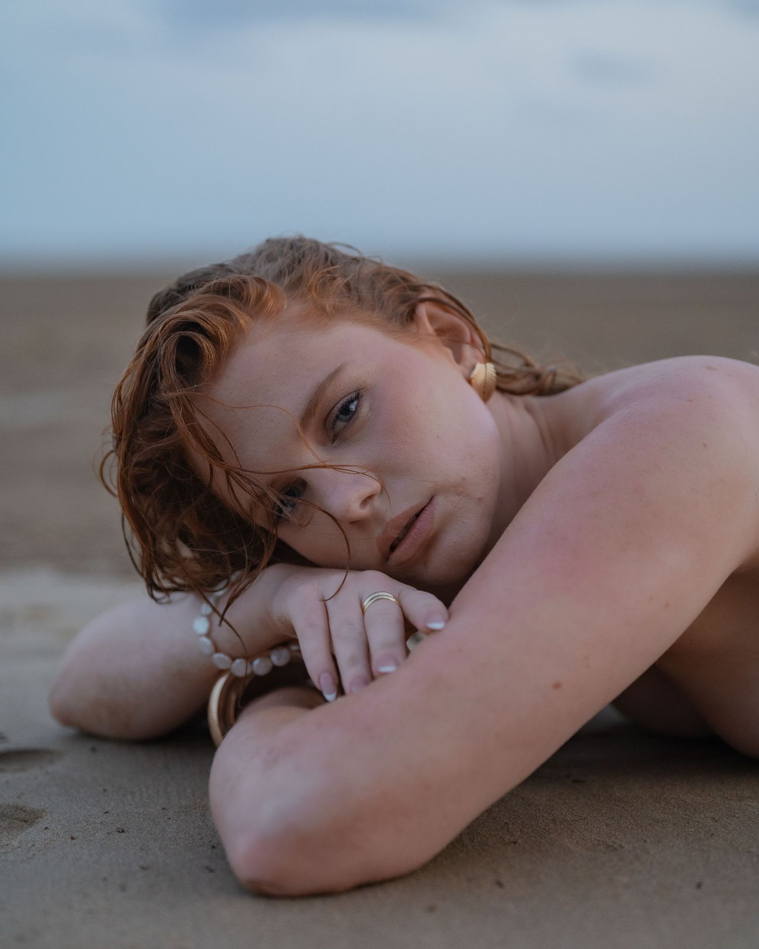 Red-haired woman with wet hair resting her head on her arm while lying on a sandy beach at dusk.