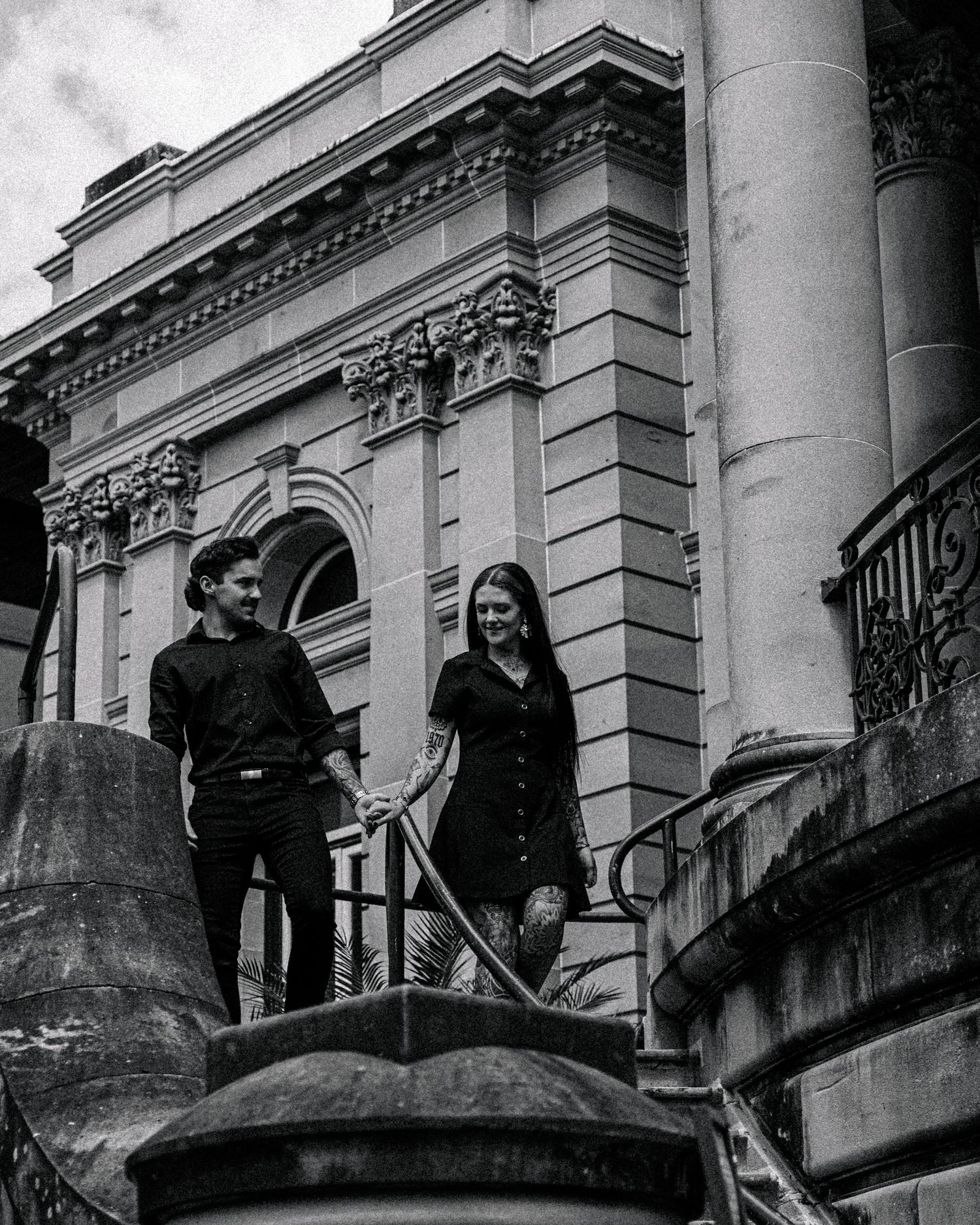 Black and white photo of a couple in dark clothing holding hands while walking down stone steps outside a grand columned building.