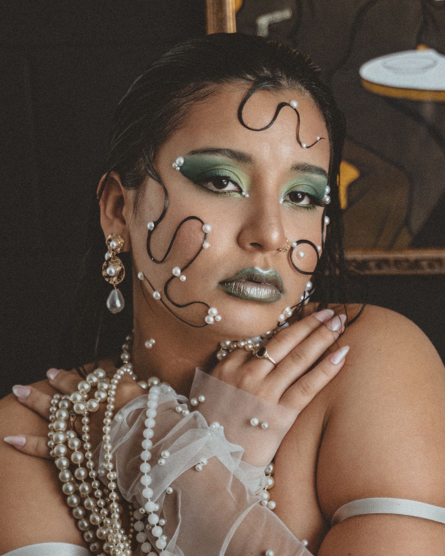 Close-up portrait of a woman with green eyeshadow, pearl-embellished wire face decor, layered pearl necklaces and sheer gloves.