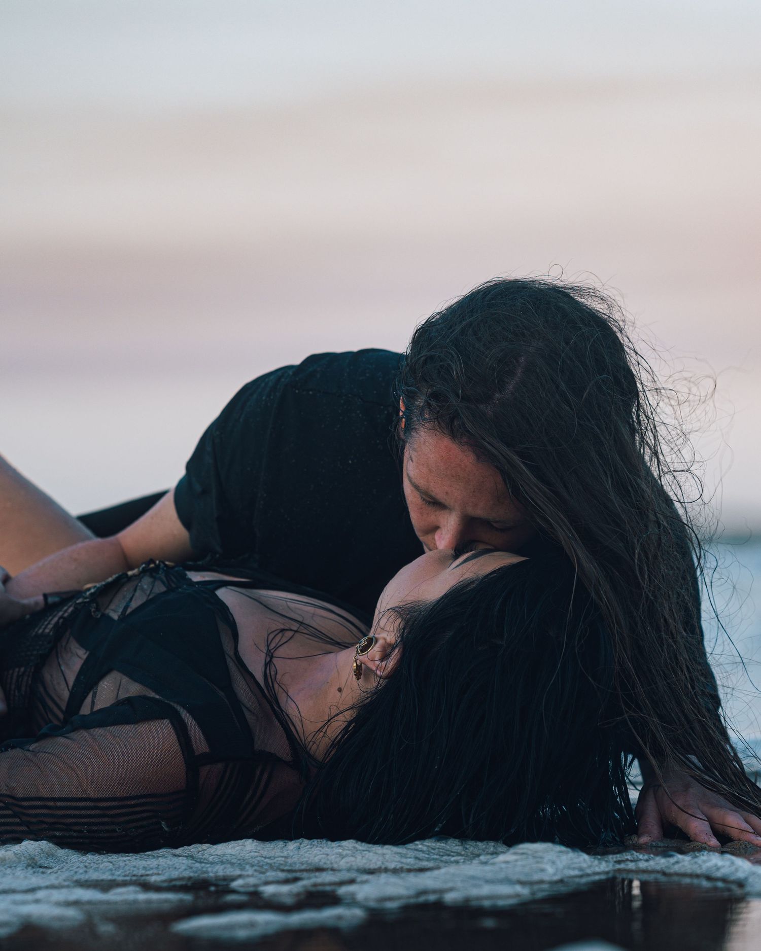 Couple in dark clothing lie close in shallow beach water at dusk, sharing a kiss with windswept hair.