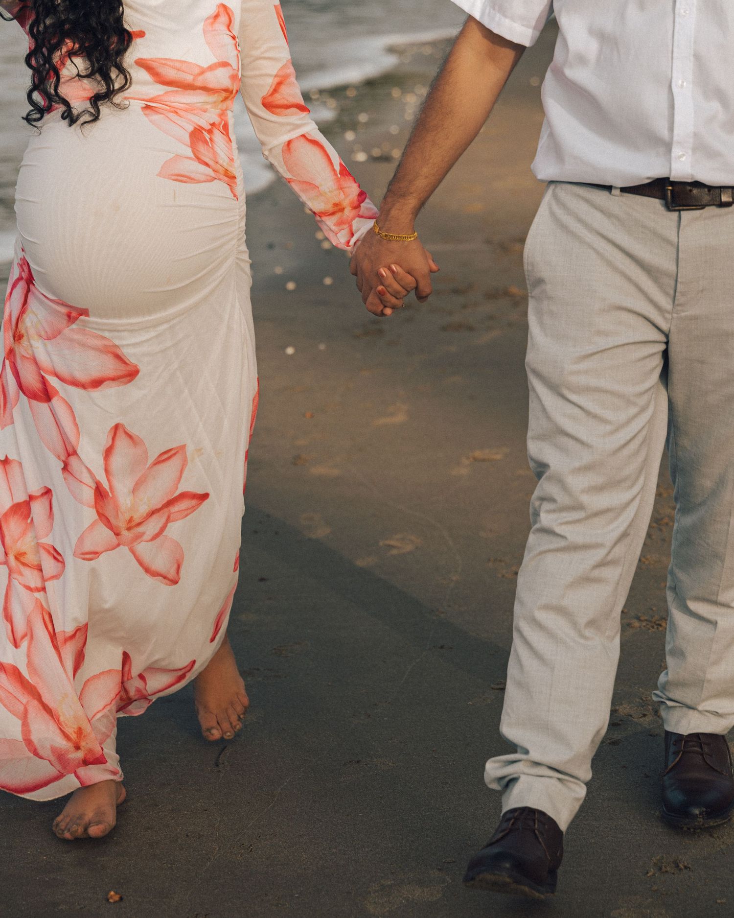 Barefoot pregnant woman in a floral dress holds hands with her partner in khakis and brown shoes while walking along the beach.