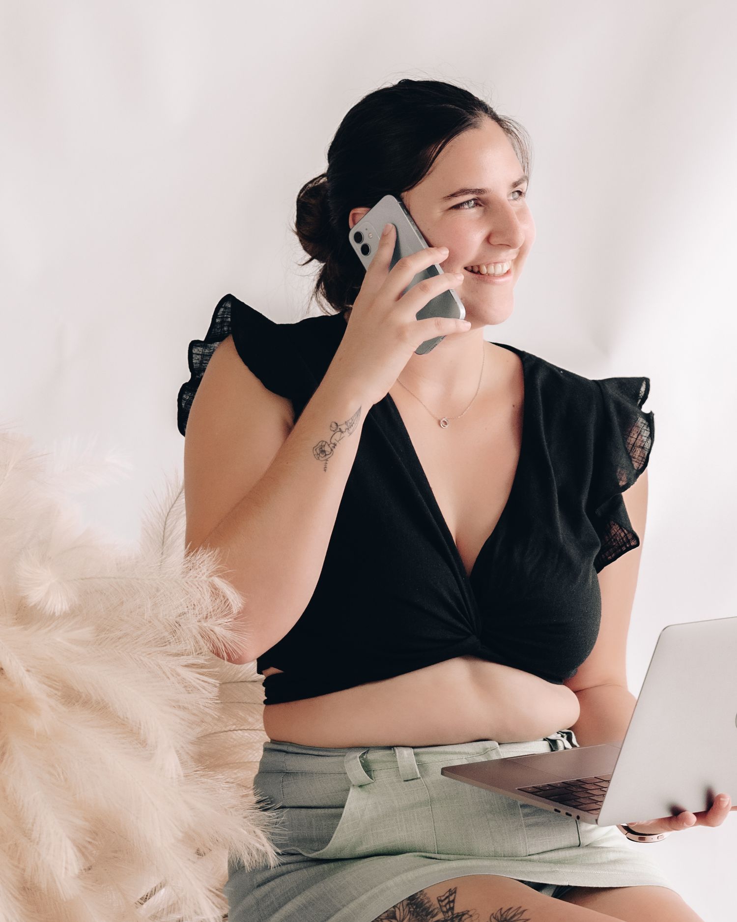 A smiling woman in a black ruffled top talks on her phone while holding a laptop in a bright studio.
