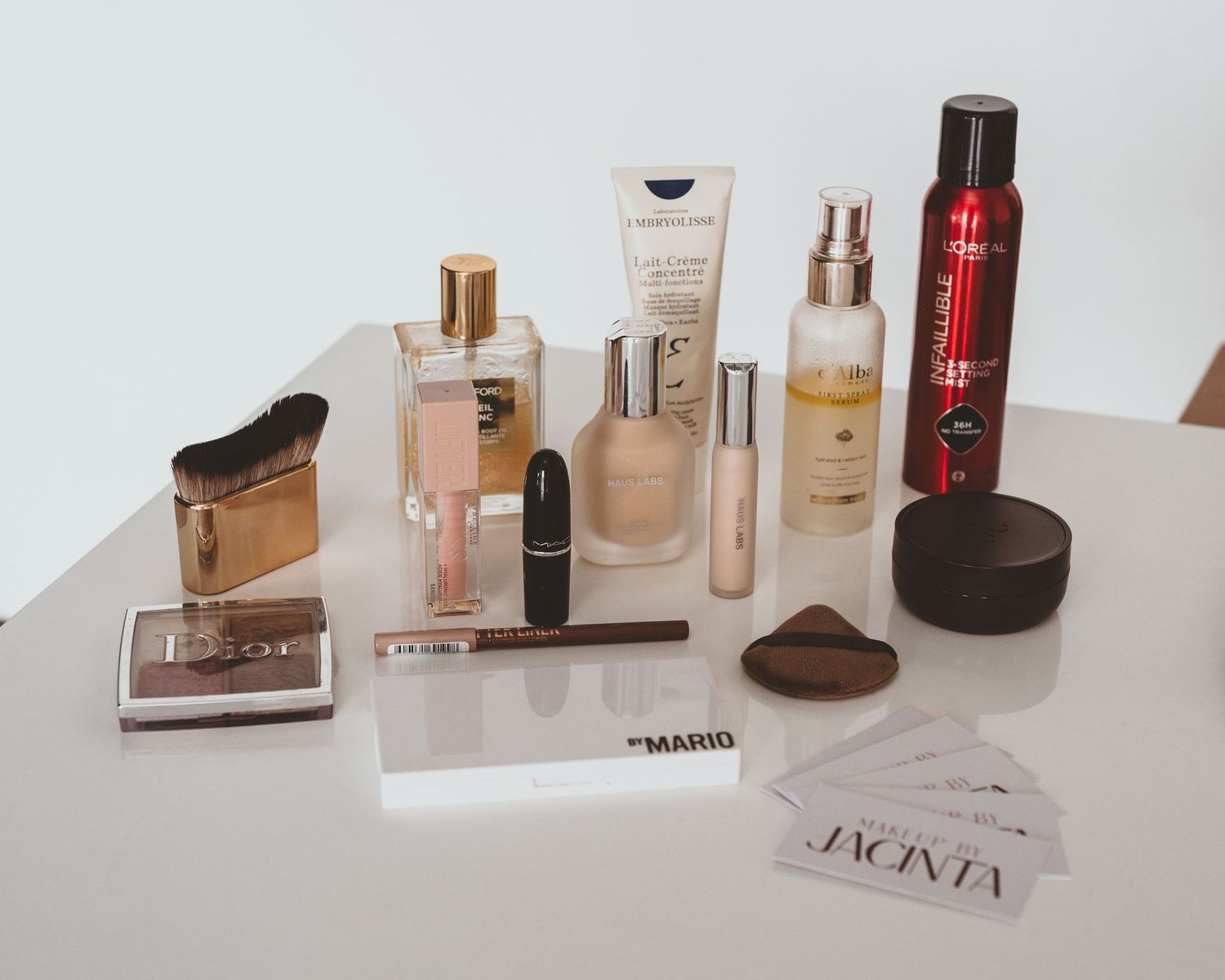 Assortment of makeup products, brushes, fragrance and Makeup by Jacinta business cards arranged on a white table.