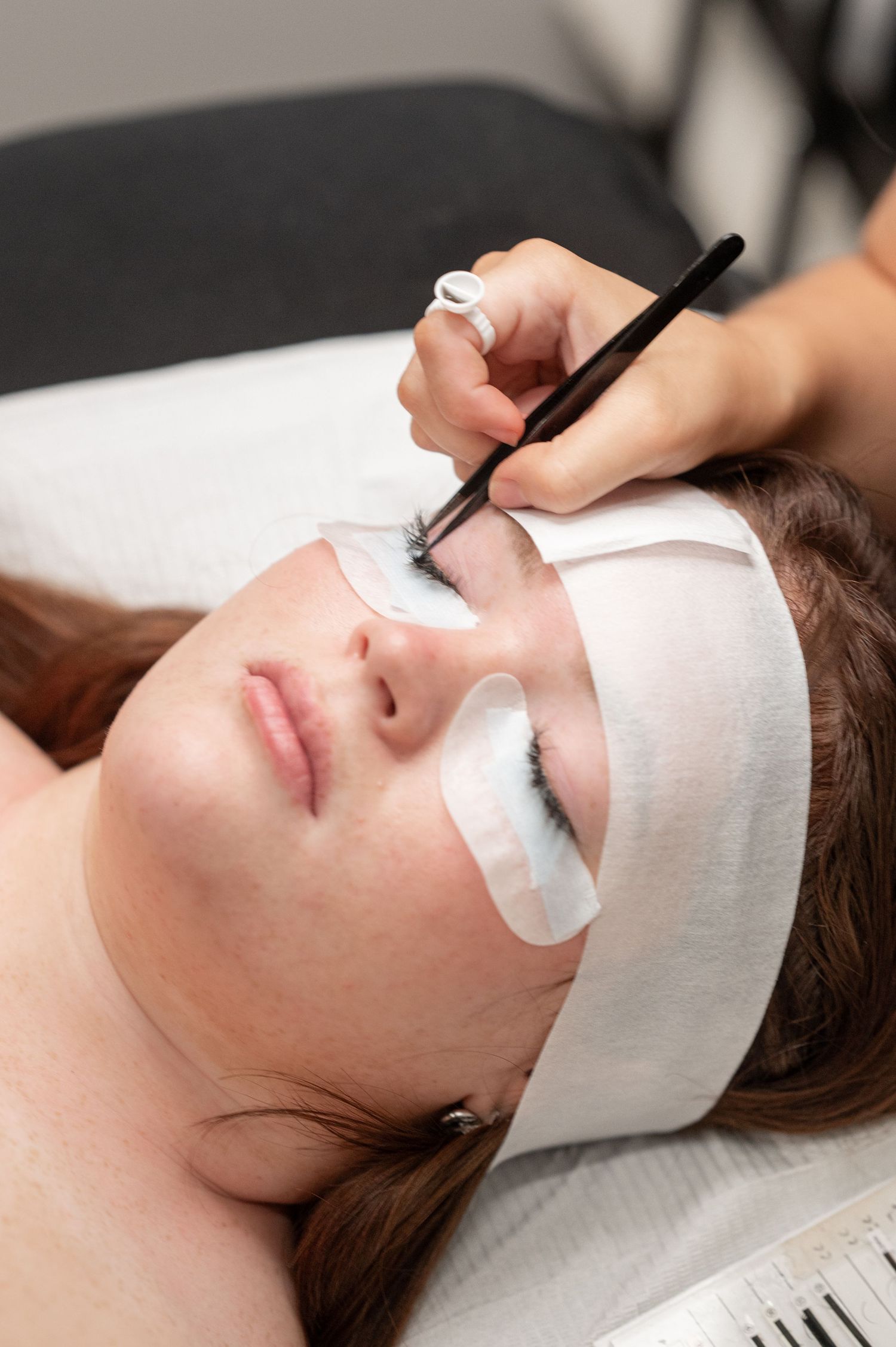 Esthetician applying lash extensions with a tool to a woman lying down with under-eye gel pads and a white headband.
