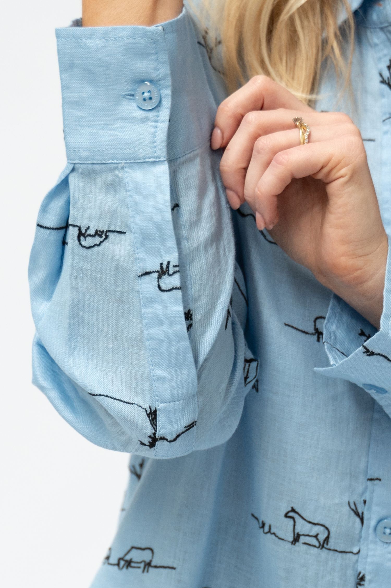 Close-up of a light blue linen shirt cuff and sleeve with black embroidered horse and tree motifs, as a woman adjusts her sleeve.
