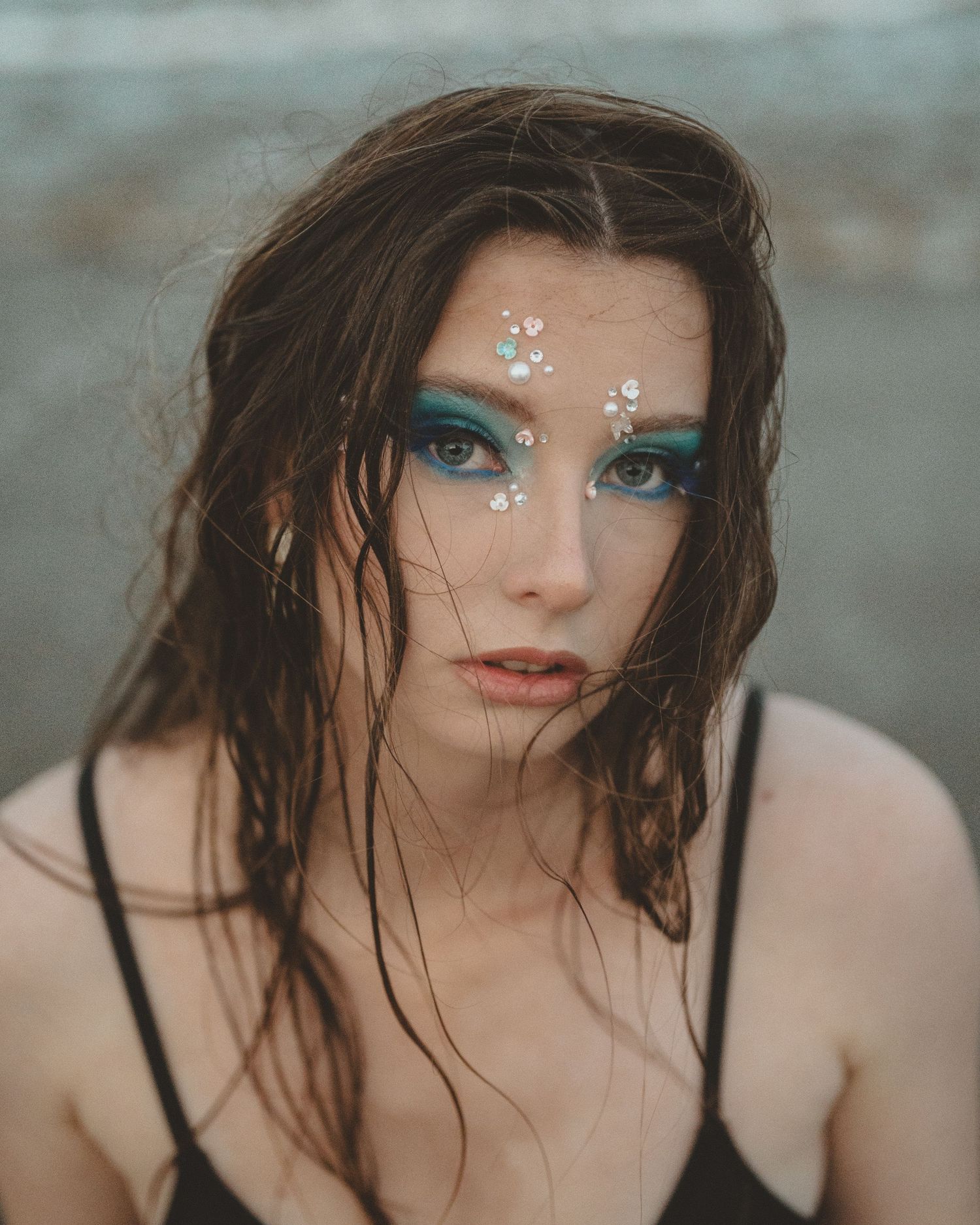 Mermaid-inspired portrait of a woman with wet tousled hair, teal eye makeup and pearl face gems standing by the ocean at dusk.