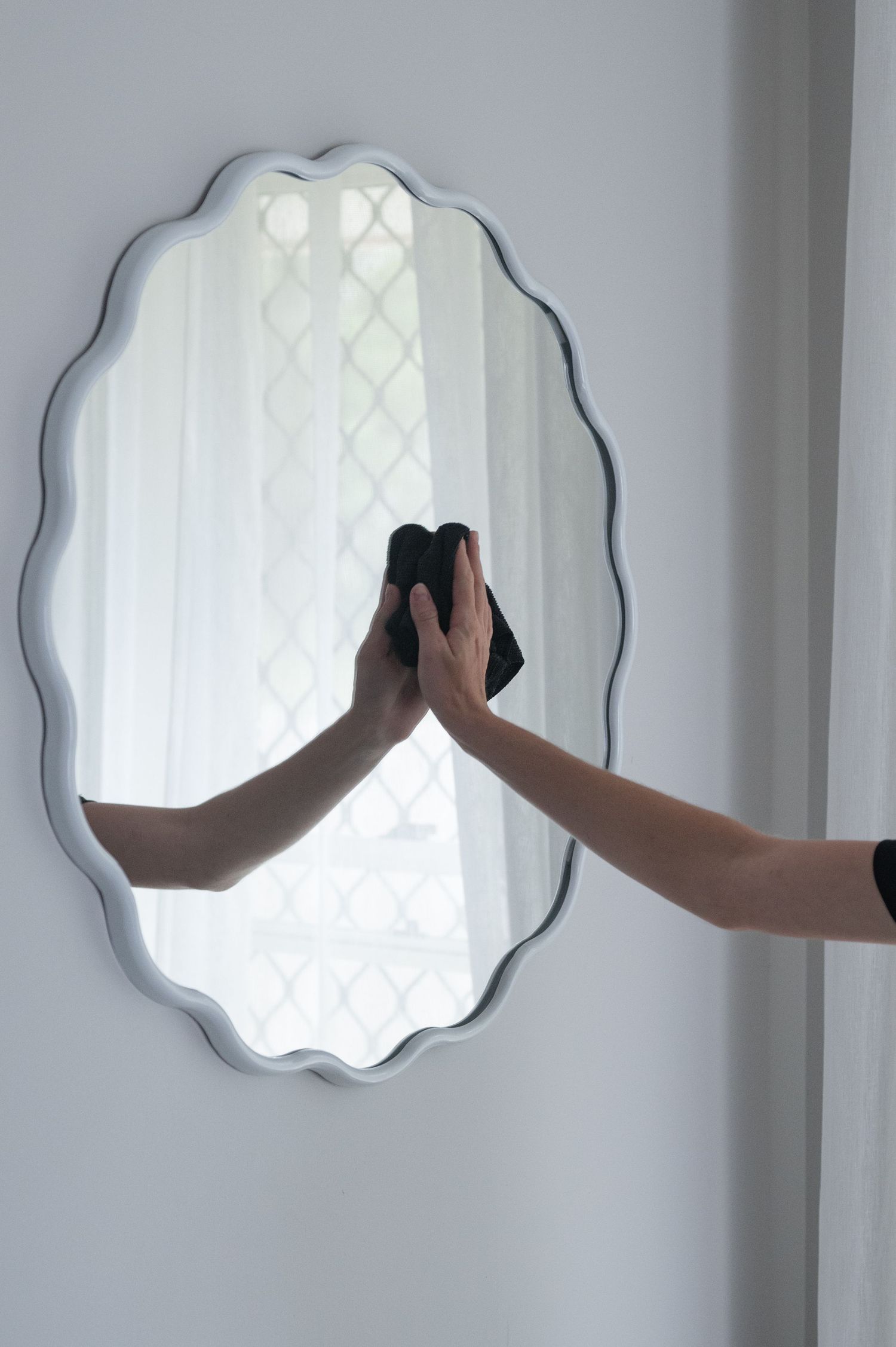 Person wiping a wavy white-framed oval mirror with a black microfiber cloth in a bright, clean room.