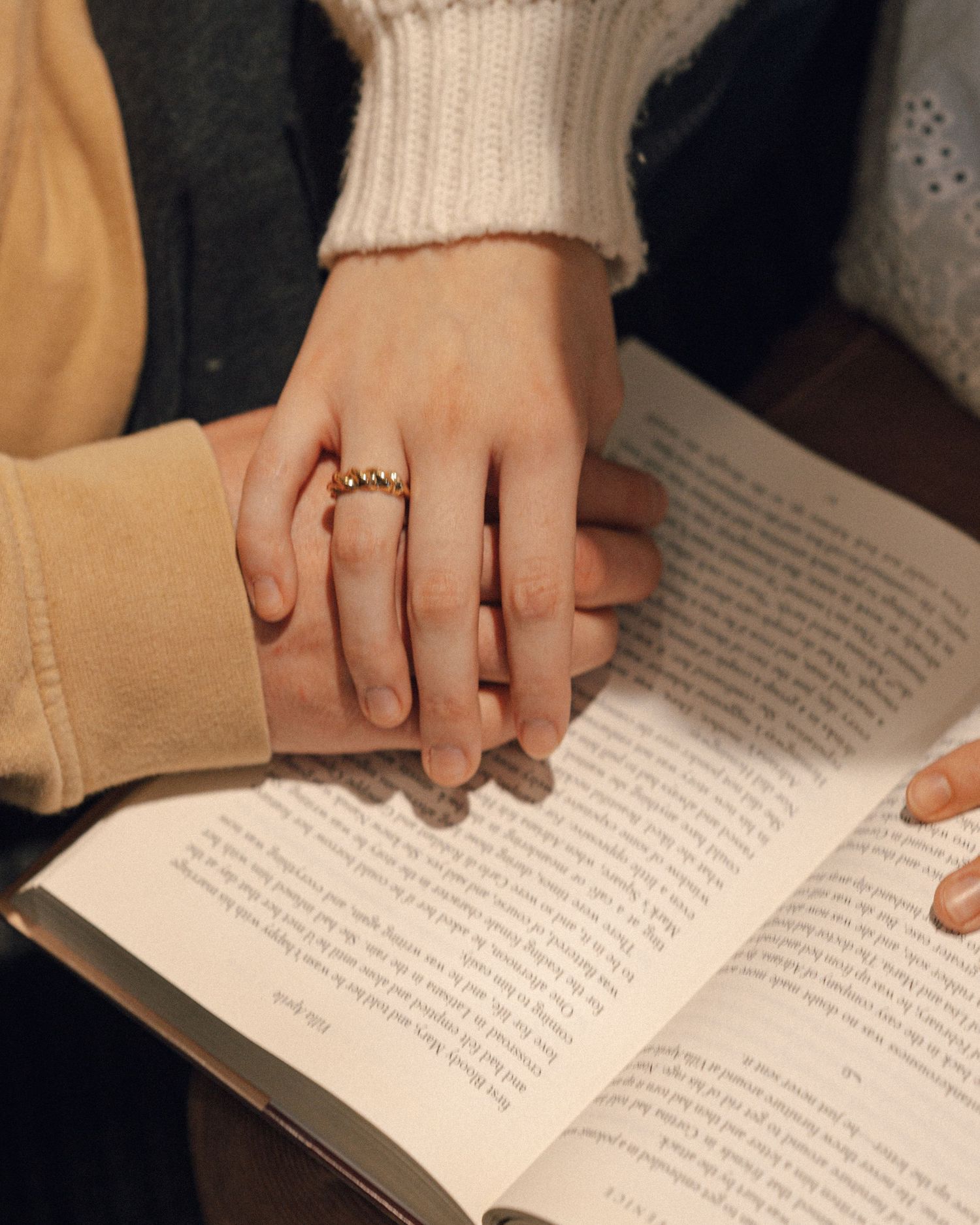 Close-up of two hands resting together on an open book, one wearing a gold ring, in a cozy reading moment.