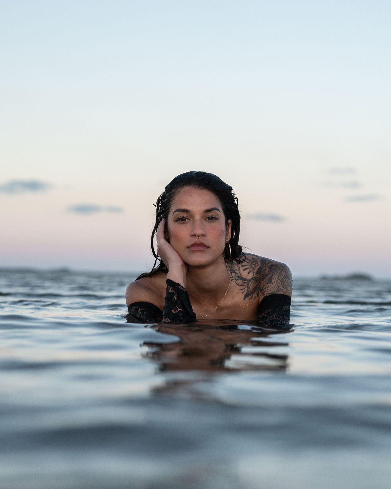 Tattooed woman with wet dark hair rests her chin on her hand, partly submerged in calm water at sunset.