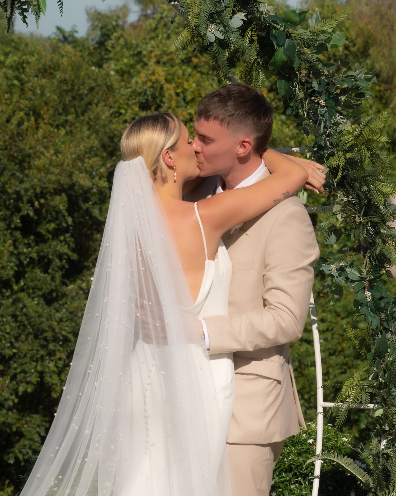 Bride in a pearl-detailed veil and groom in a beige suit share a kiss in front of a greenery wedding arch.