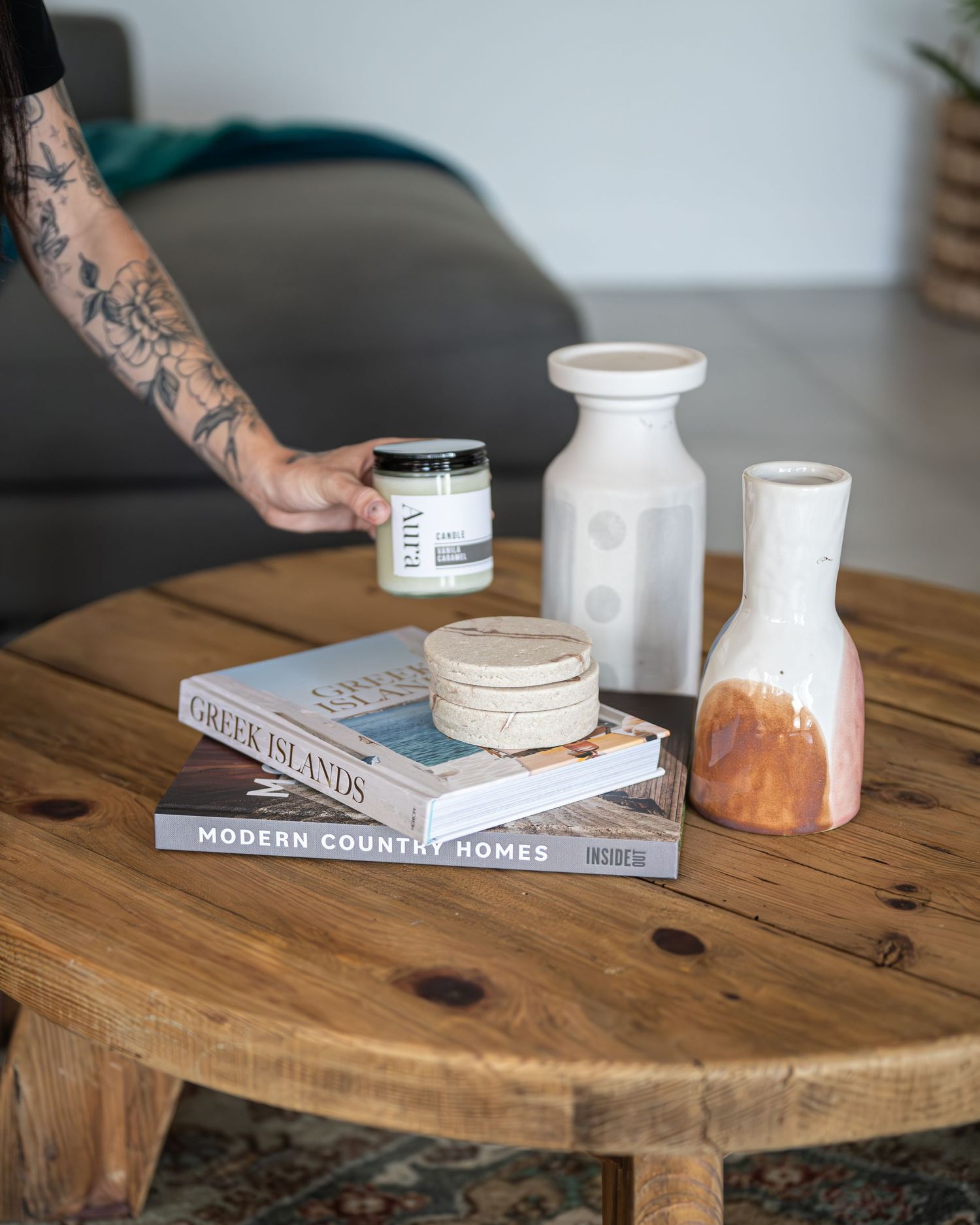 Hand placing an Aura candle on a round rustic wooden coffee table with books, stone coasters and ceramic vases.