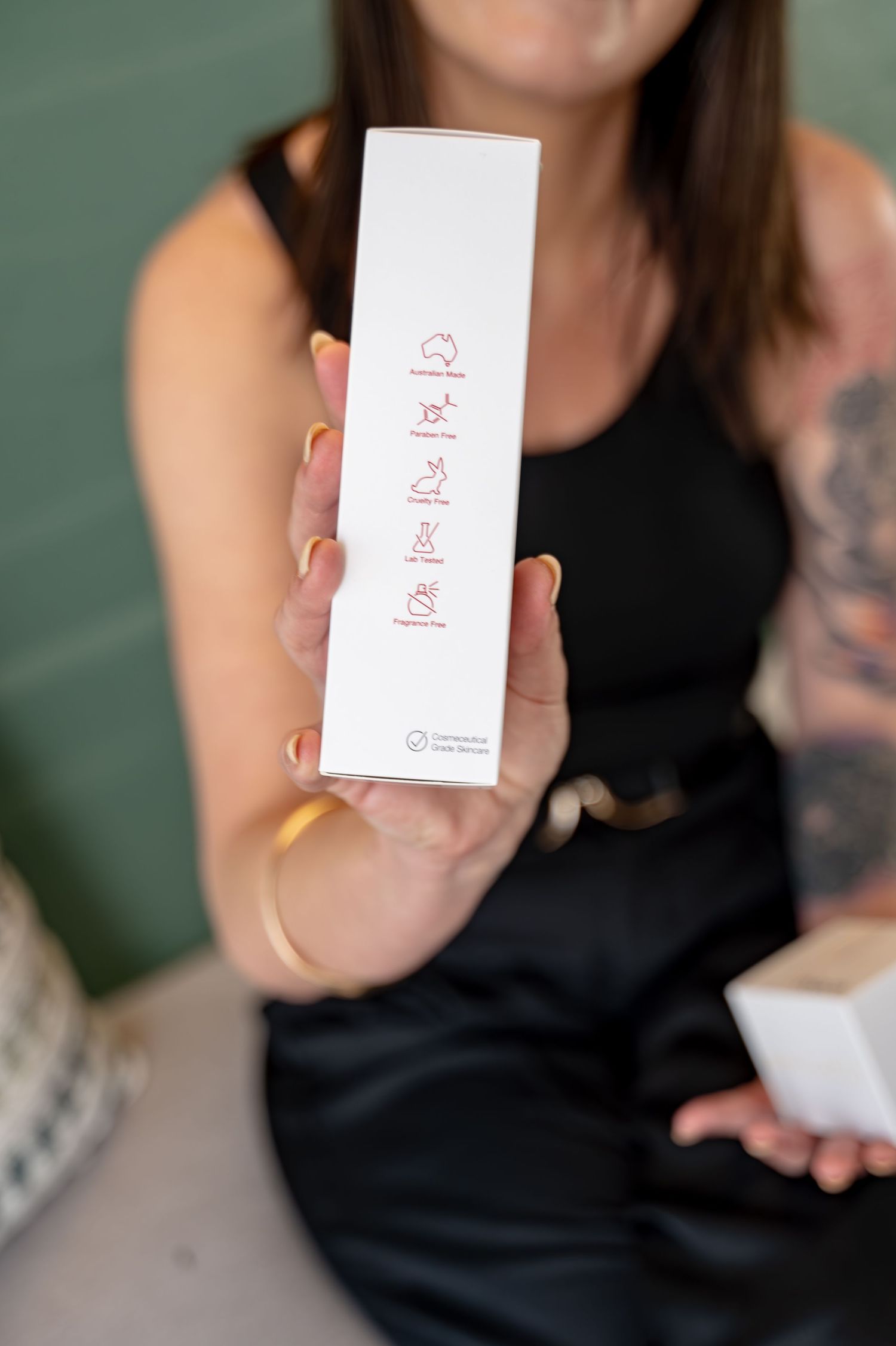 Woman holds up a white skincare box showing red icons for product claims while seated, with a tattooed arm and gold bracelet visible.