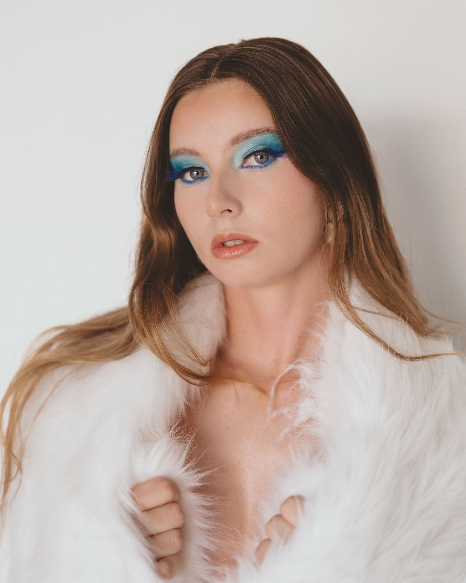 Woman with vibrant blue eyeshadow and long ombre hair wrapped in a fluffy white faux fur coat posing against a white studio background.