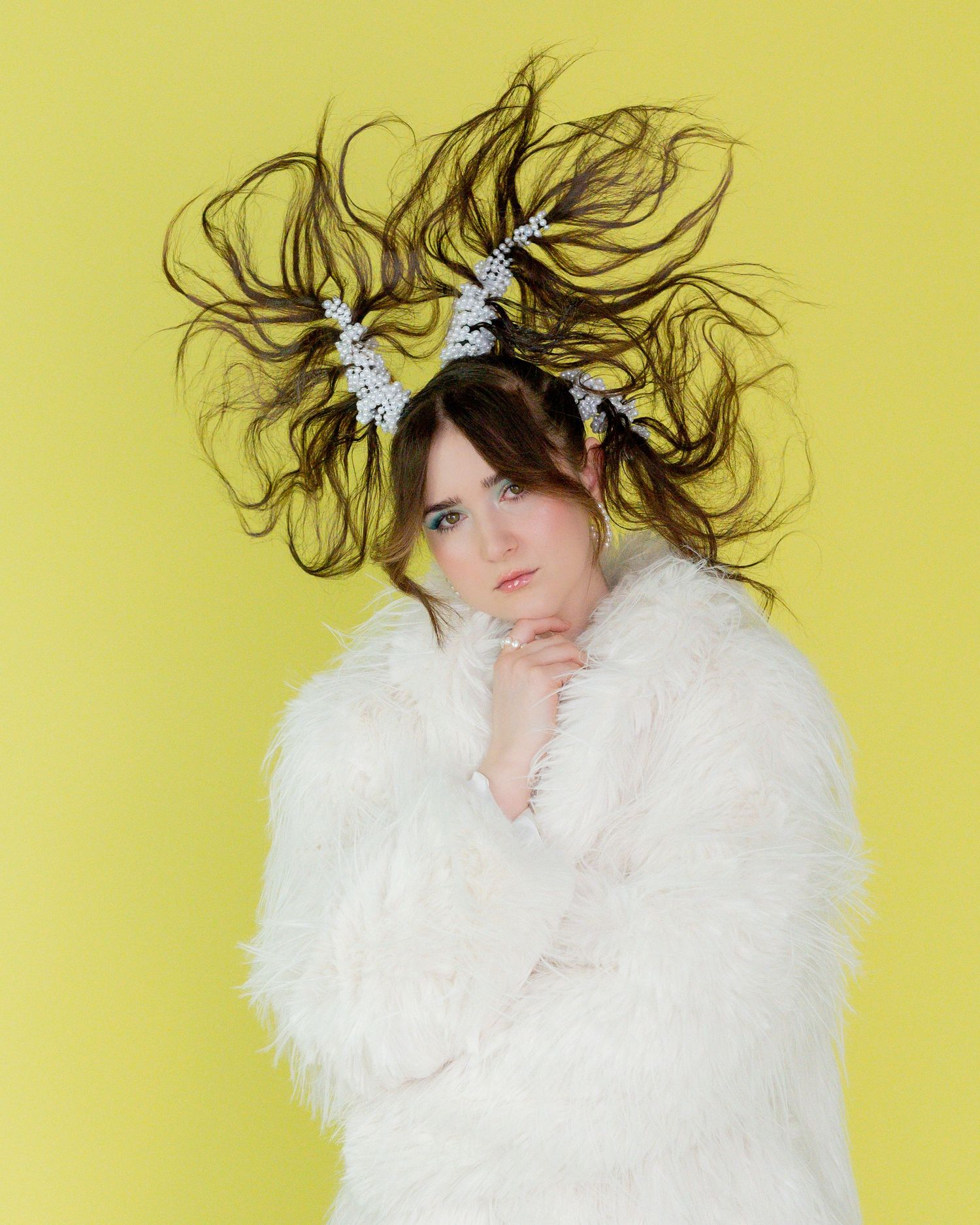 Model in a fluffy white fur coat with wild, windswept hair and white floral hair accessories posing against a bright yellow background.