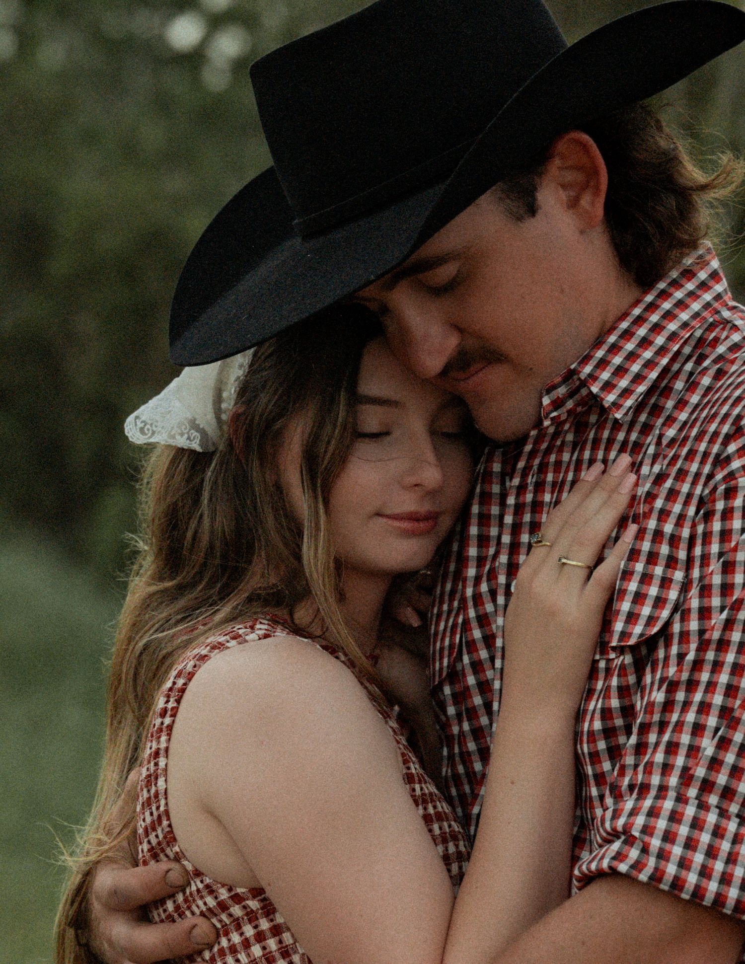 A woman rests her head on the chest of a mustached man in a black cowboy hat and plaid shirt in a peaceful outdoor embrace.