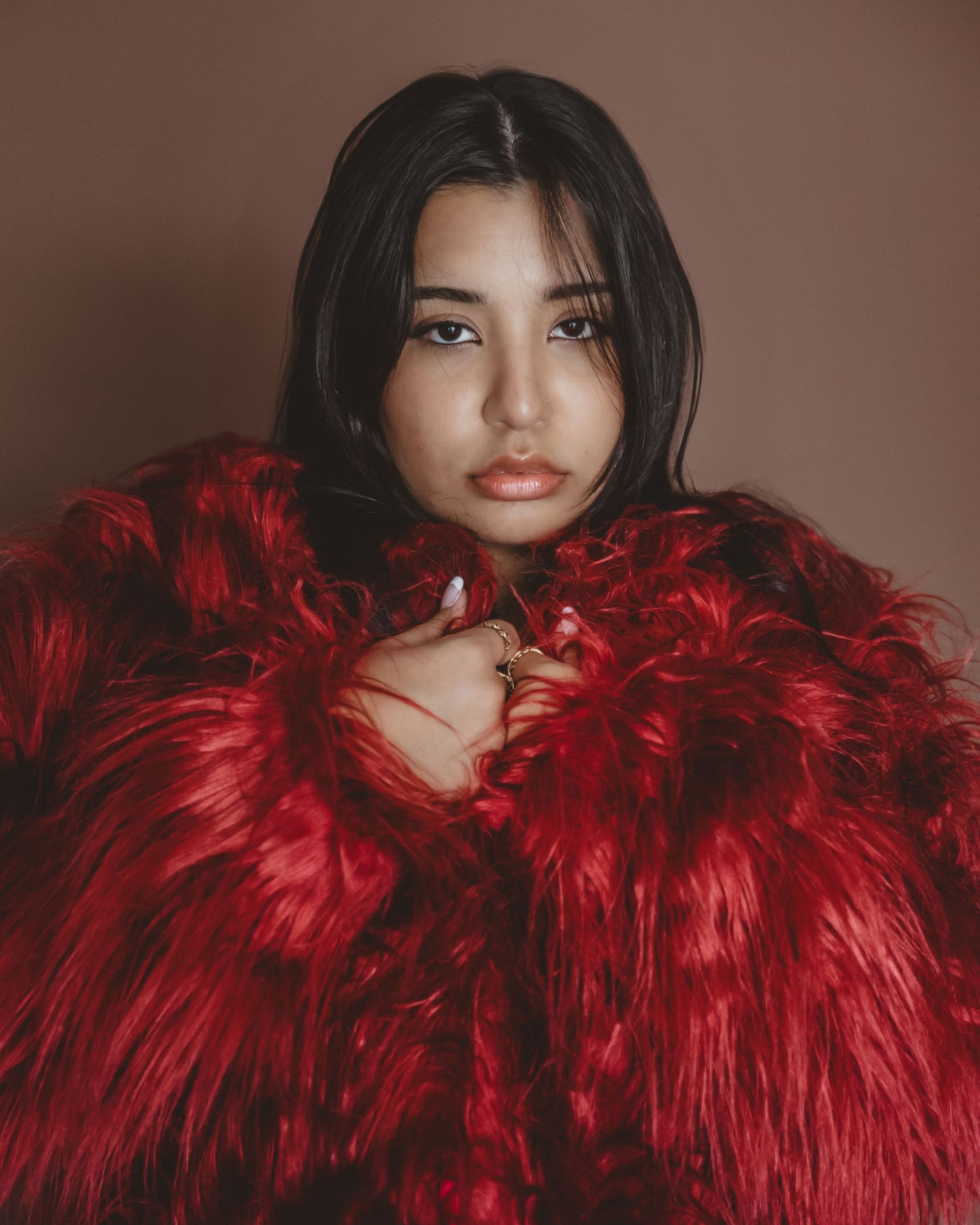 Woman with long dark hair wearing an oversized red faux fur coat, posing against a muted brown studio background.