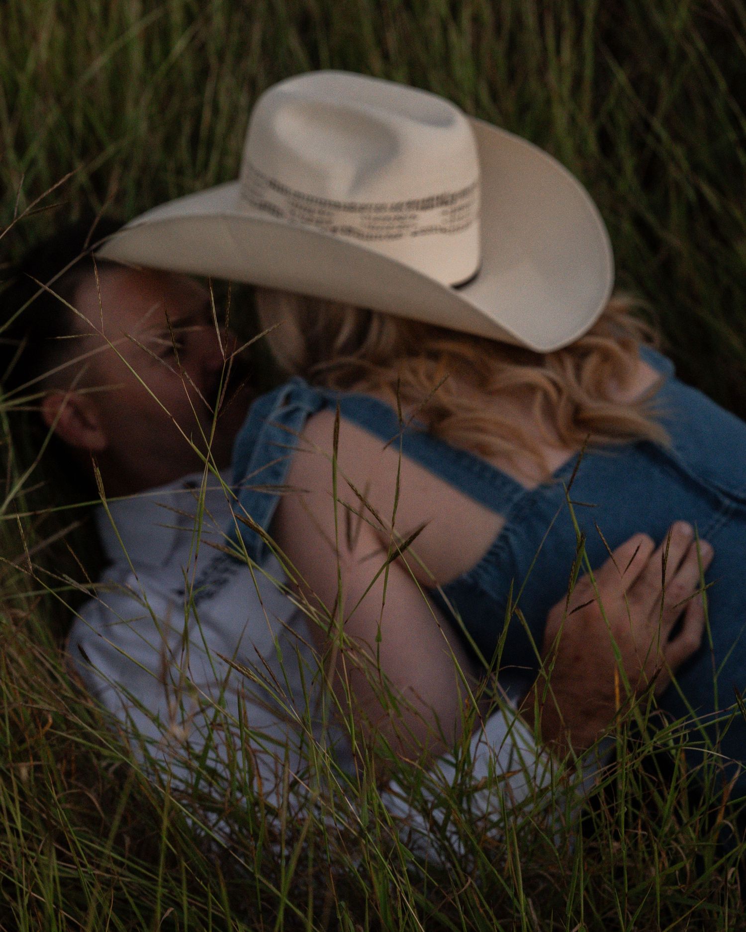A close-up of a couple in cowboy hats embracing in tall grass, the woman in a denim dress with wavy blonde hair.