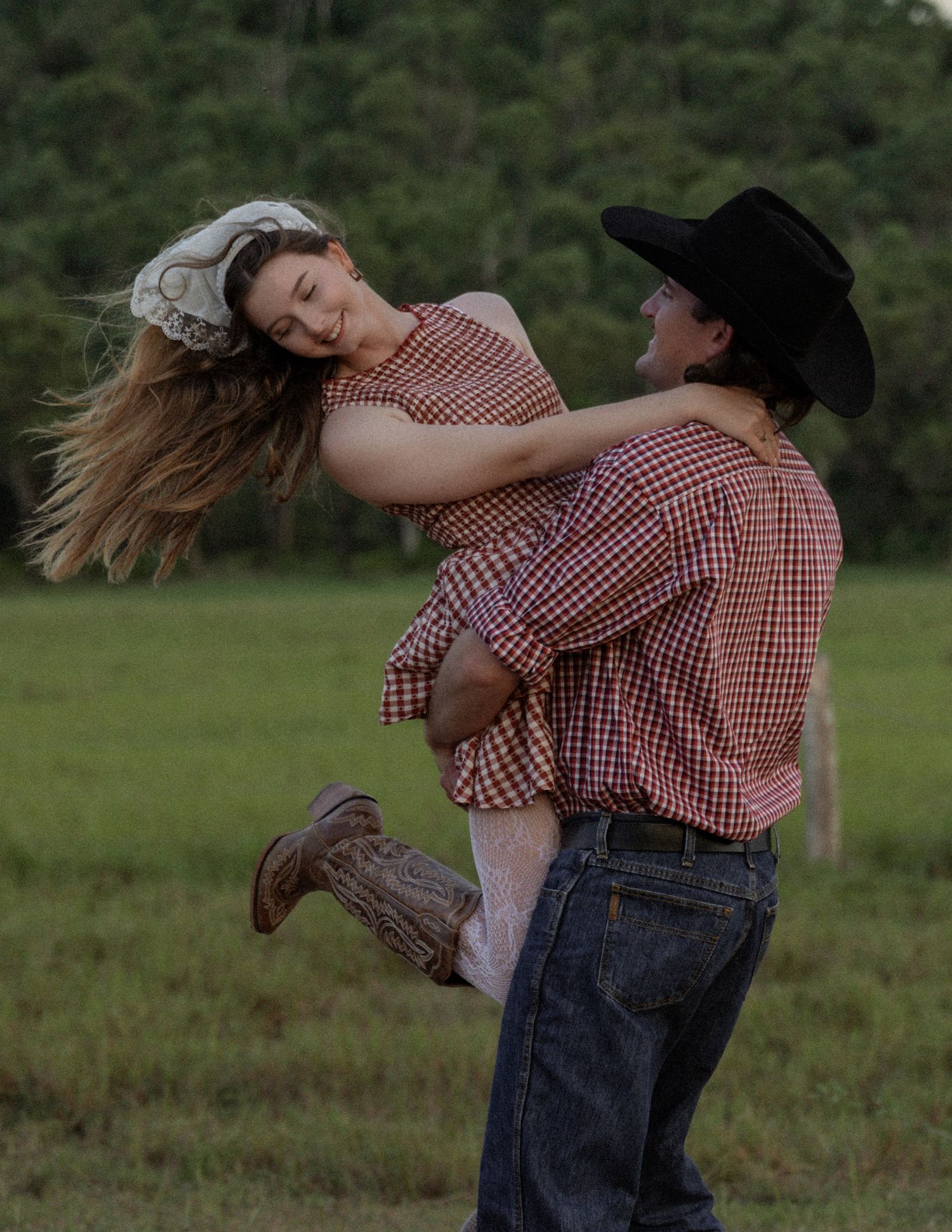 Woman in a gingham dress and head scarf laughs as a cowboy in a black hat swings her through the air on a green pasture.