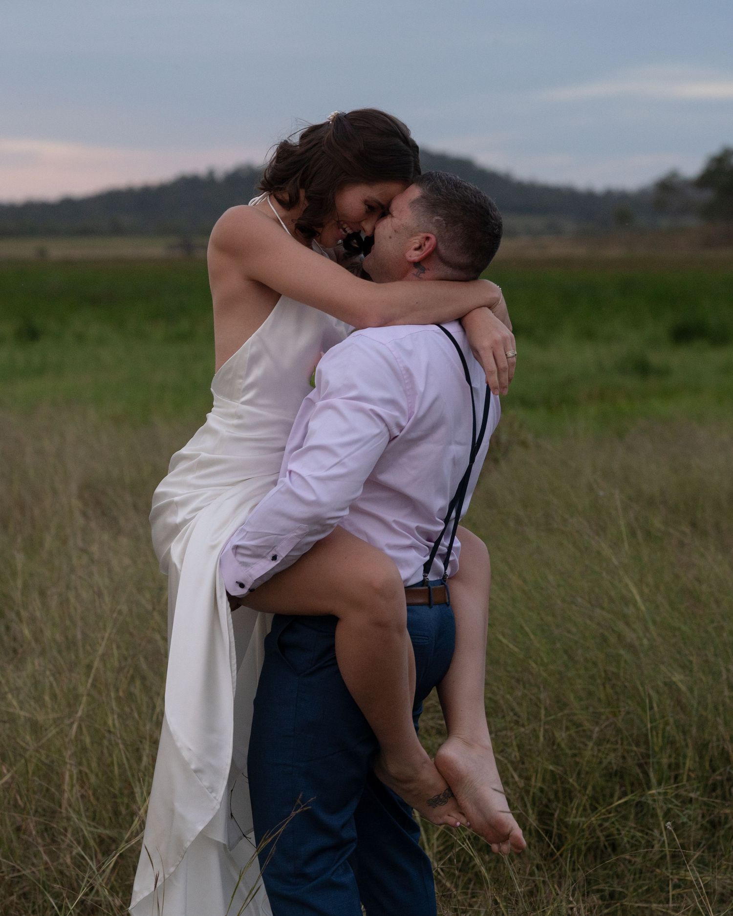 Groom in suspenders lifts his bride in a white gown as they embrace in a grassy field at dusk.