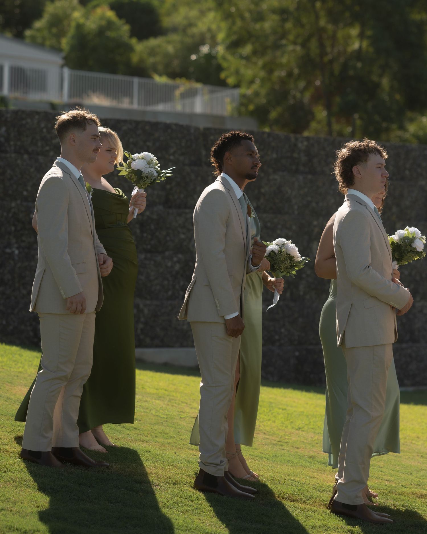 Groomsmen in beige suits and bridesmaids in olive green dresses holding white bouquets stand on a sunlit lawn.