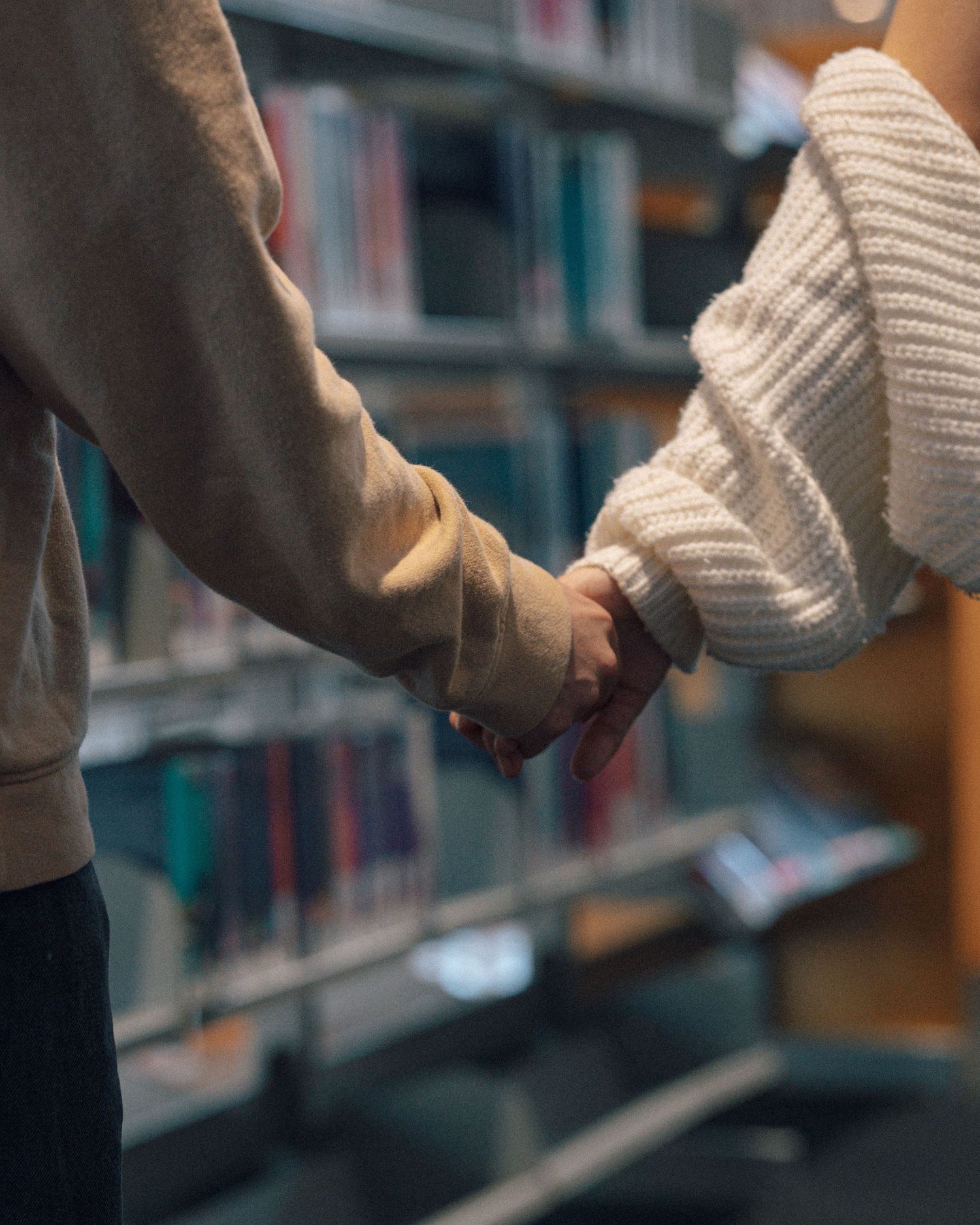 Close-up of a couple holding hands in a library aisle, wearing cozy knit and beige sweaters beside tall bookshelves.