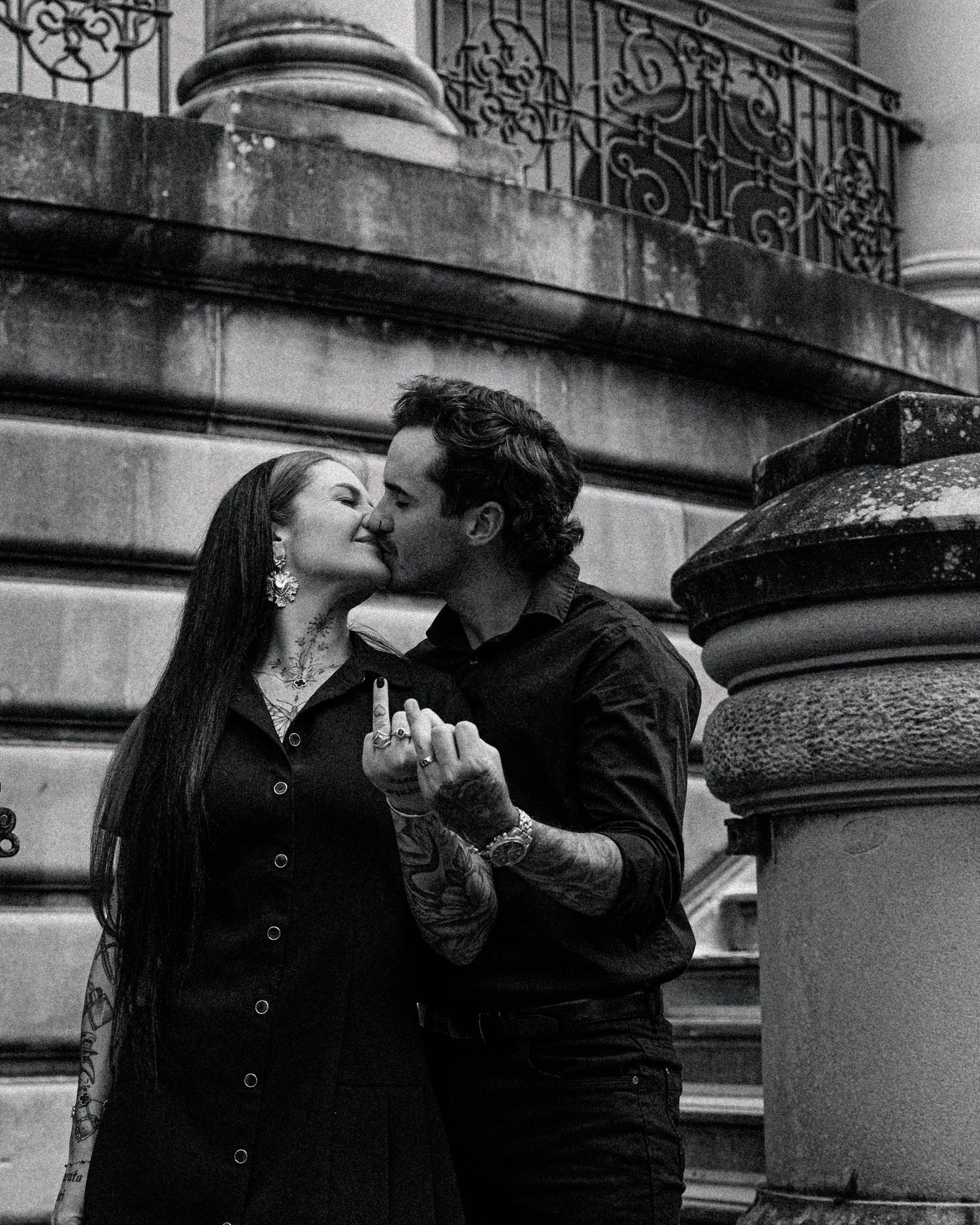 Black and white image of a tattooed couple kissing on stone steps as the woman shows off her engagement ring.