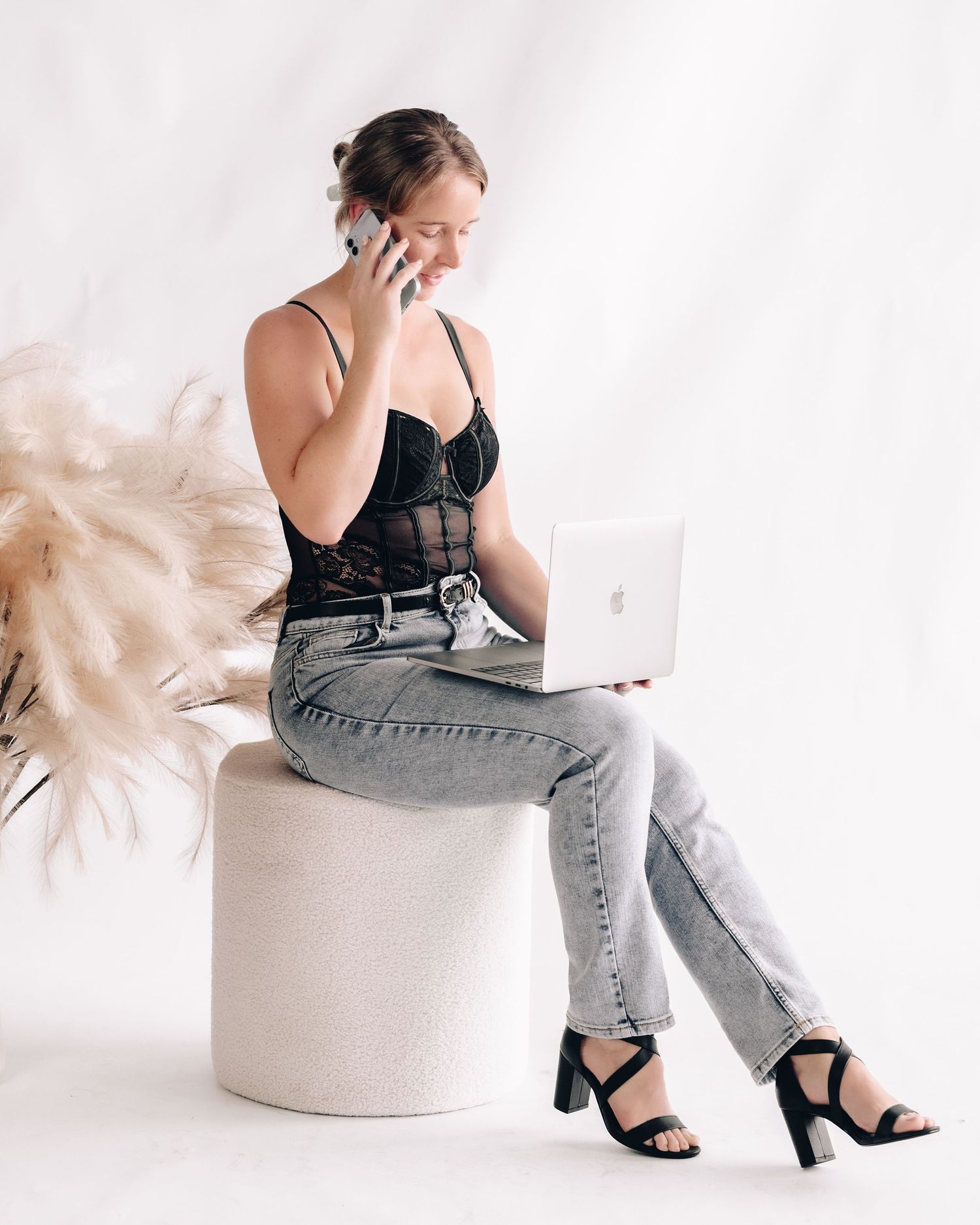 A woman in a black lace bodysuit and gray jeans sits on a white stool, talking on the phone with a laptop on her lap.