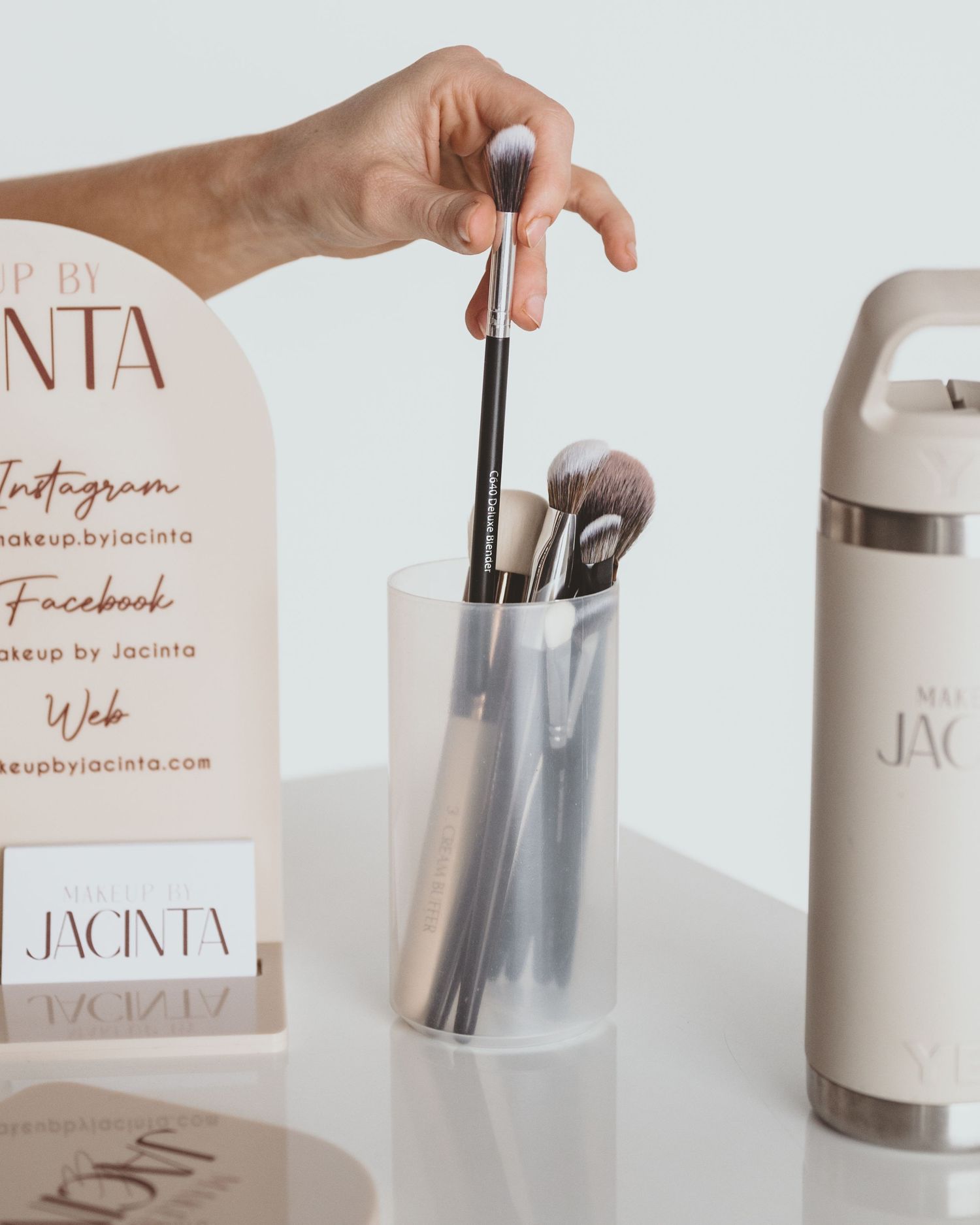 Hand pulls a makeup brush from a clear holder next to a Makeup by Jacinta social media sign and a cream water bottle.