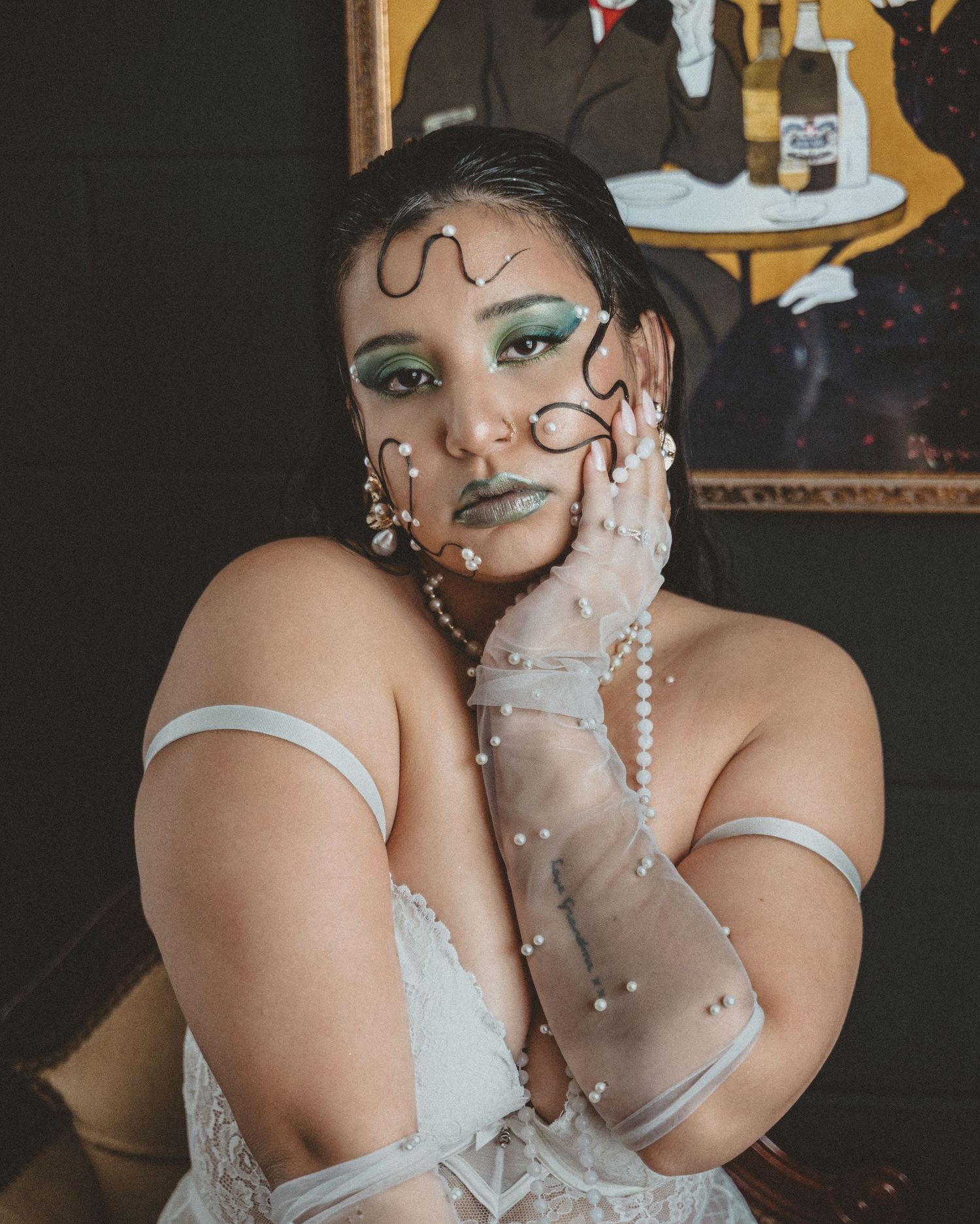 Woman rests her face on her hand, wearing avant-garde green makeup, pearl face accents and white lace lingerie near a framed painting.
