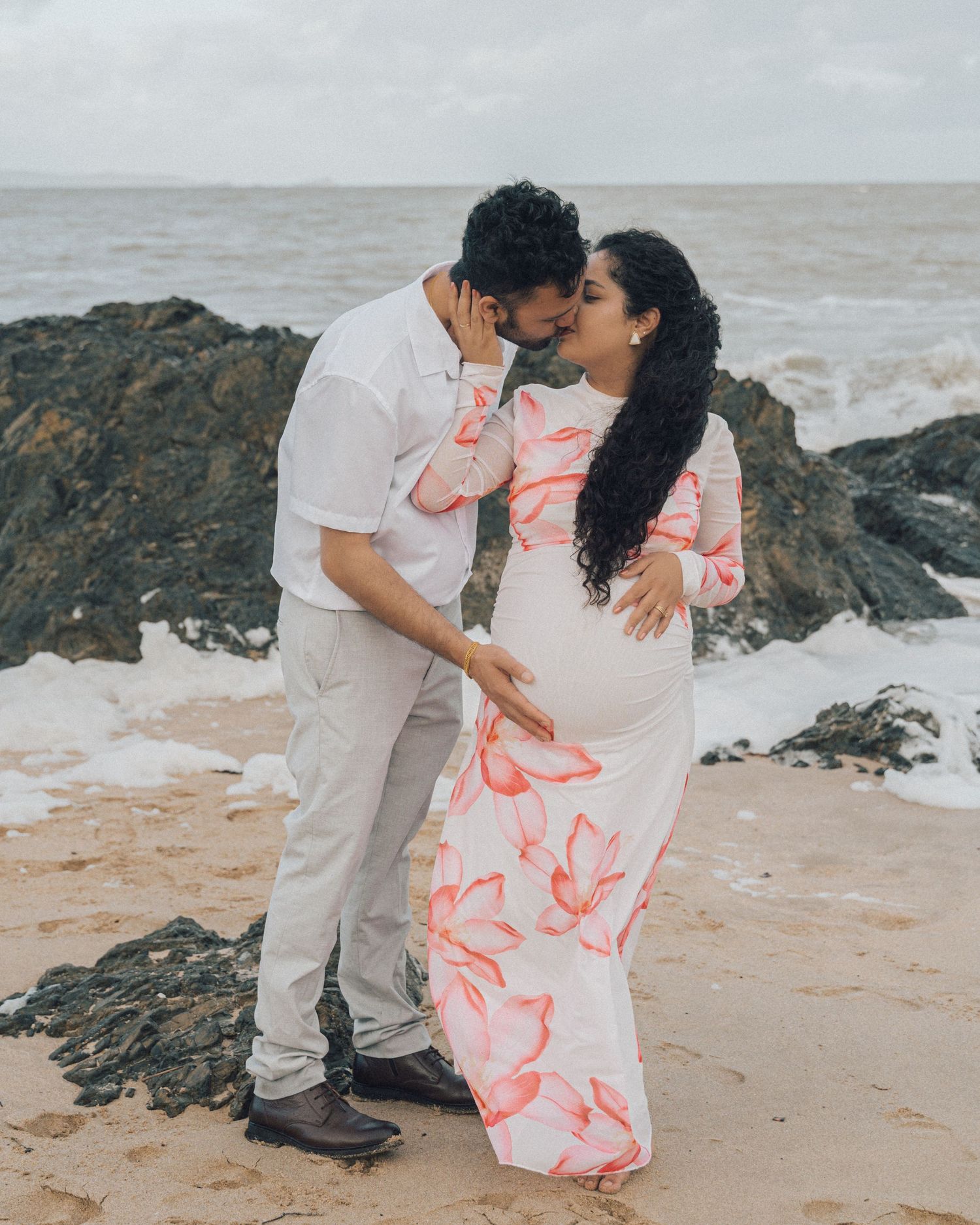 Expecting couple shares a kiss on a rocky beach, the woman in a pink floral maternity dress and the man in a white shirt.