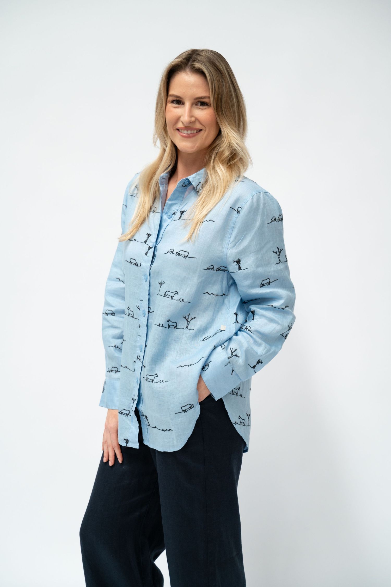 A smiling woman wears a light blue linen button-up shirt with an embroidered horse and landscape print and navy wide-leg pants.