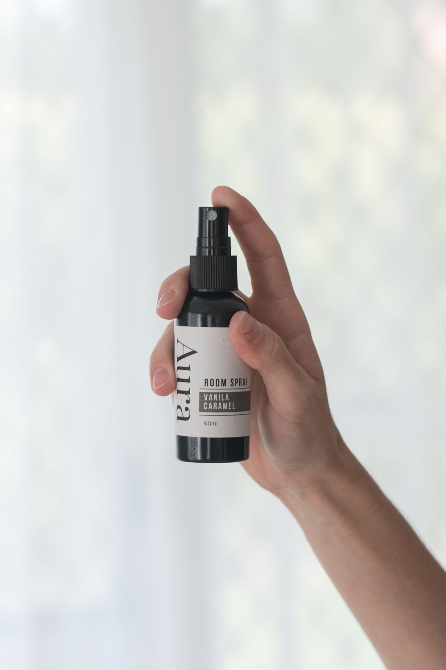 Hand holding an Aura vanilla caramel room spray bottle with a black spray top against a soft, bright background.