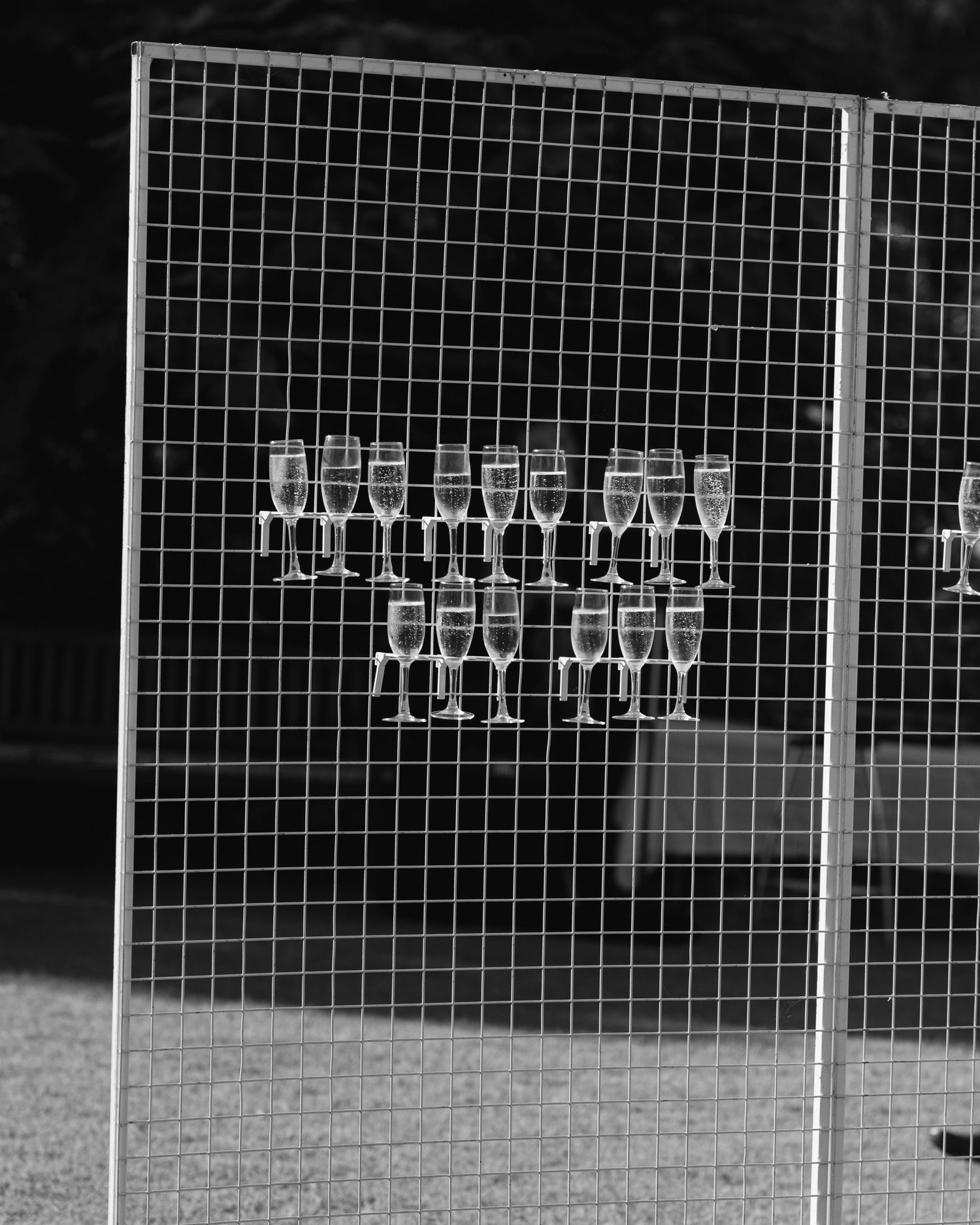 Black and white view of champagne glasses arranged on a wire mesh display wall at an outdoor wedding.