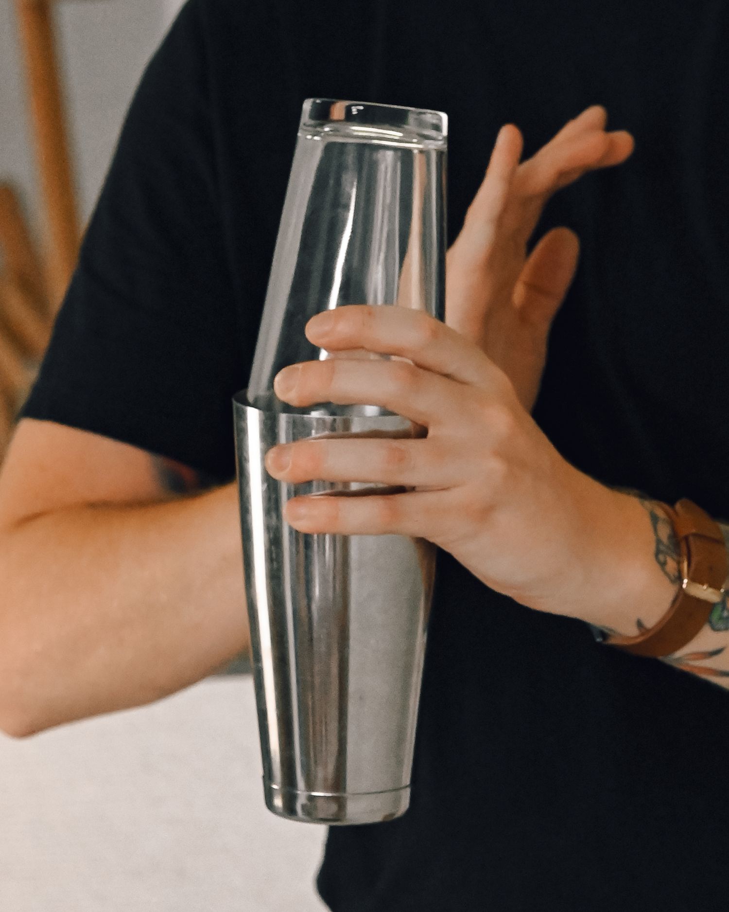 Bartender's hands hold a stainless steel and glass Boston cocktail shaker, wearing a brown leather watch and black tee.