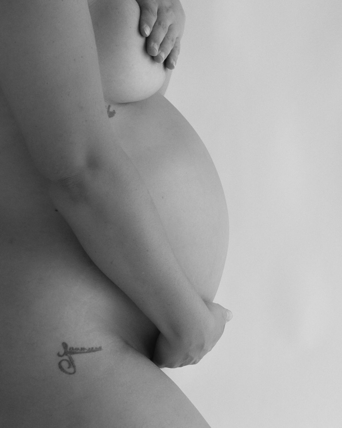 Black and white side profile of a pregnant woman cradling her bare belly with a small tattoo on her hip.
