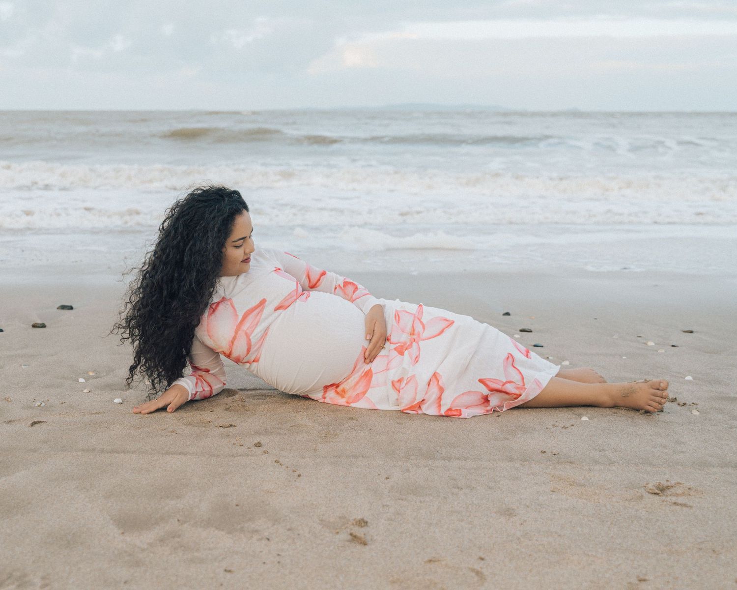 Expectant mother reclines on a quiet beach in a floral maternity dress, gazing toward the waves under a soft cloudy sky.