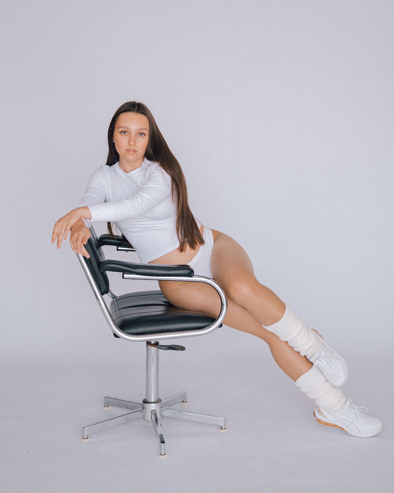Woman in a white long-sleeve top and leg warmers reclines on a black and chrome swivel chair against a soft gray backdrop.